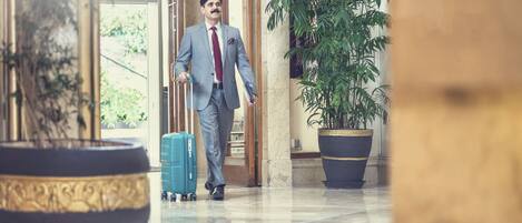A man in a suit walking with a suitcase in a hotel lobby with marble floors and large potted plants.