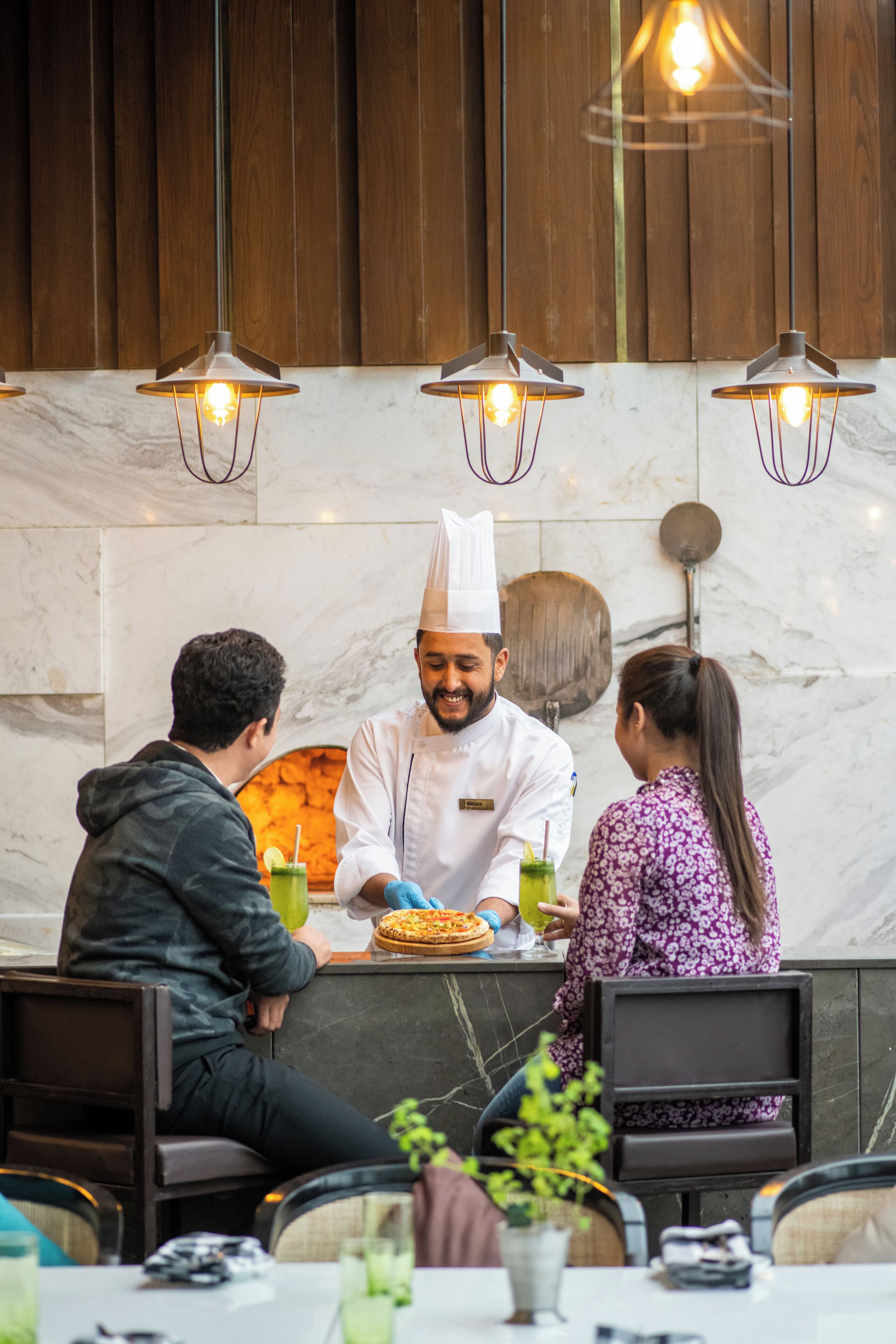 A restaurant with a marble countertop, hanging pendant lights, and a chef preparing food.