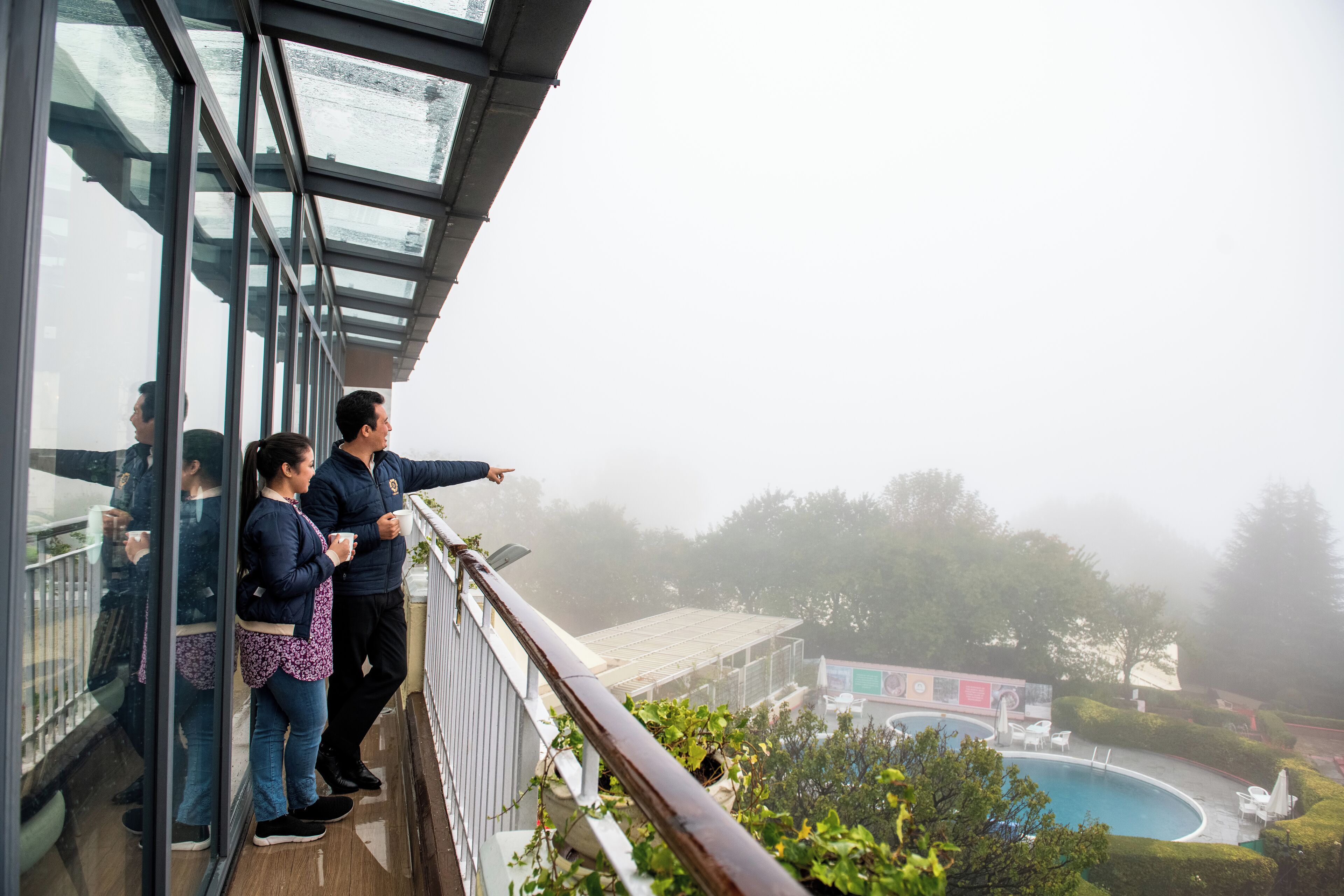A balcony with a glass railing overlooking a pool area, surrounded by greenery and a misty atmosphere.