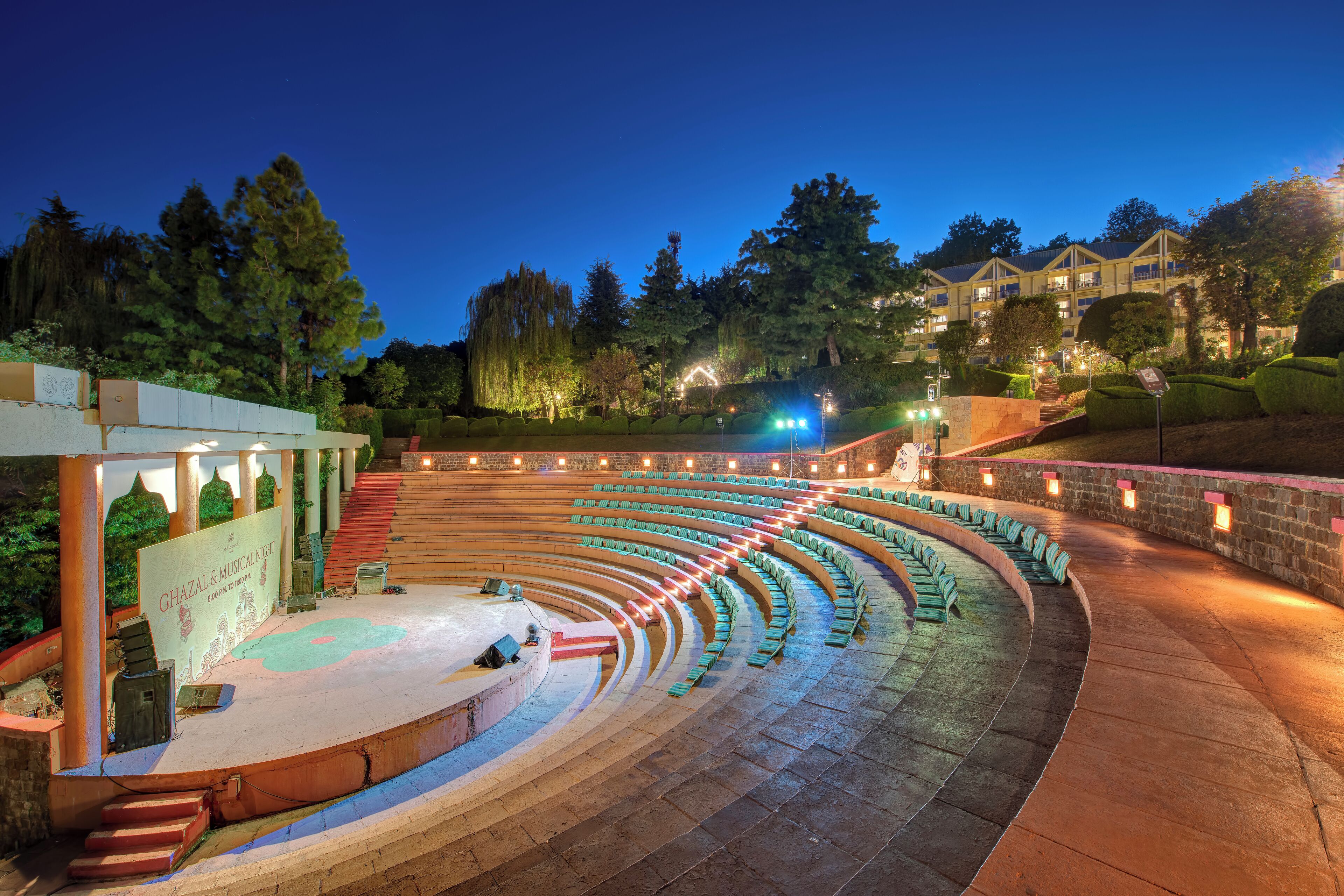 An outdoor amphitheater with tiered seating and a stage, surrounded by trees and a building in the background.