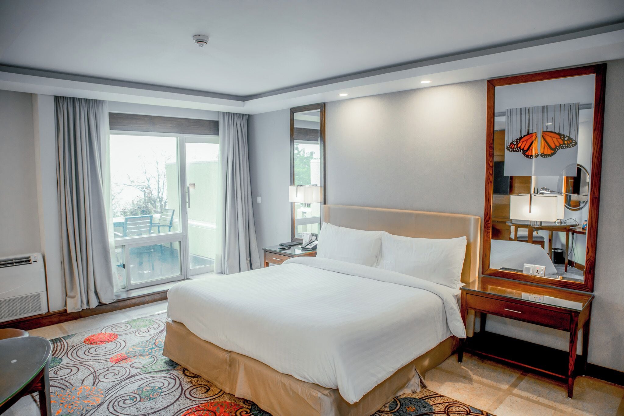 A hotel room with a large bed, a desk, a chair, a window with curtains, and a mirror.