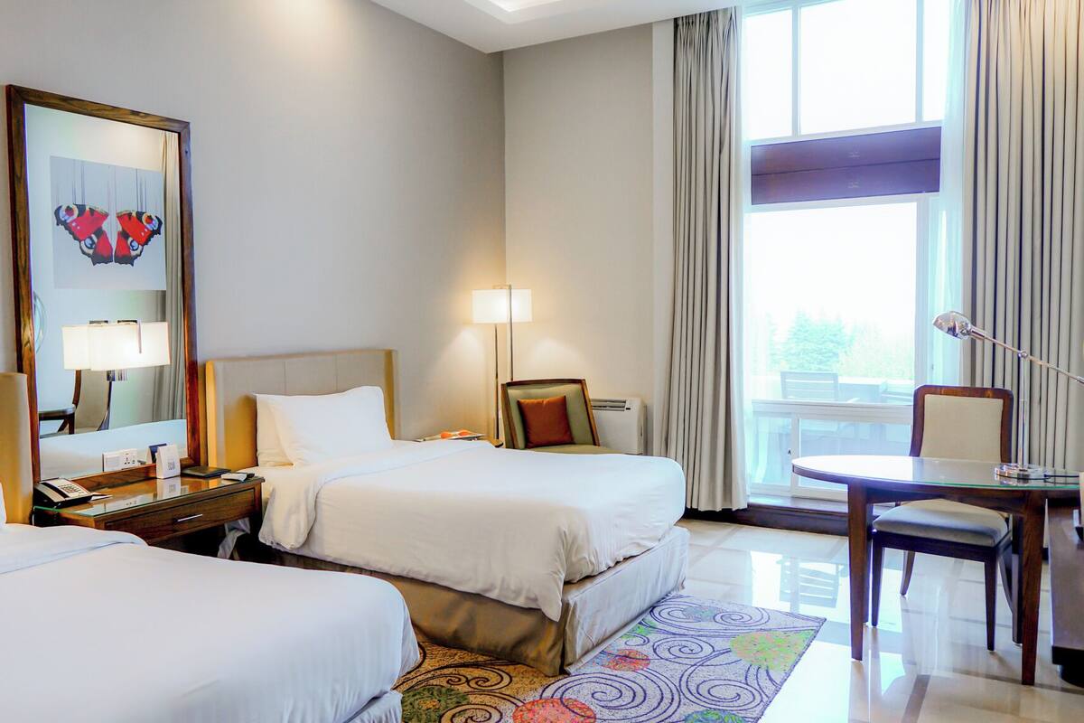 Une chambre d’hôtel avec deux lits, un bureau, une chaise, une lampe, un miroir et une fenêtre avec des rideaux.