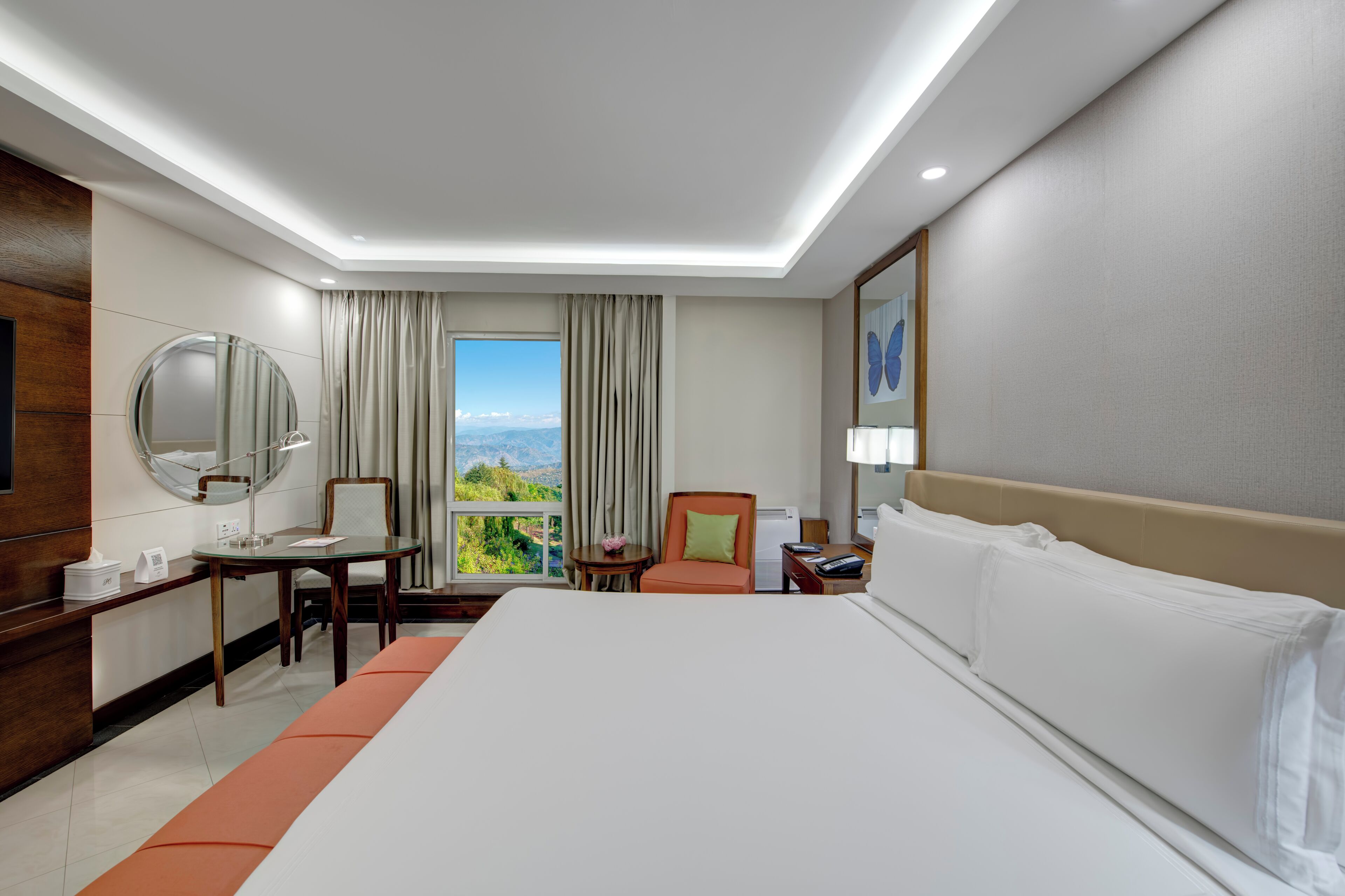 Habitación de hotel con una cama grande, un escritorio con silla, un espejo redondo, una mesita de noche y vistas al exterior.