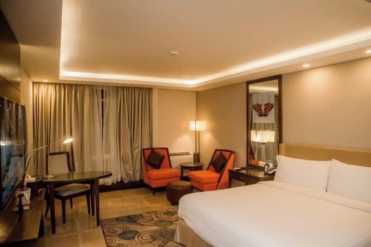 A hotel room with a bed, two chairs, a desk, a lamp, a mirror, and curtains.
