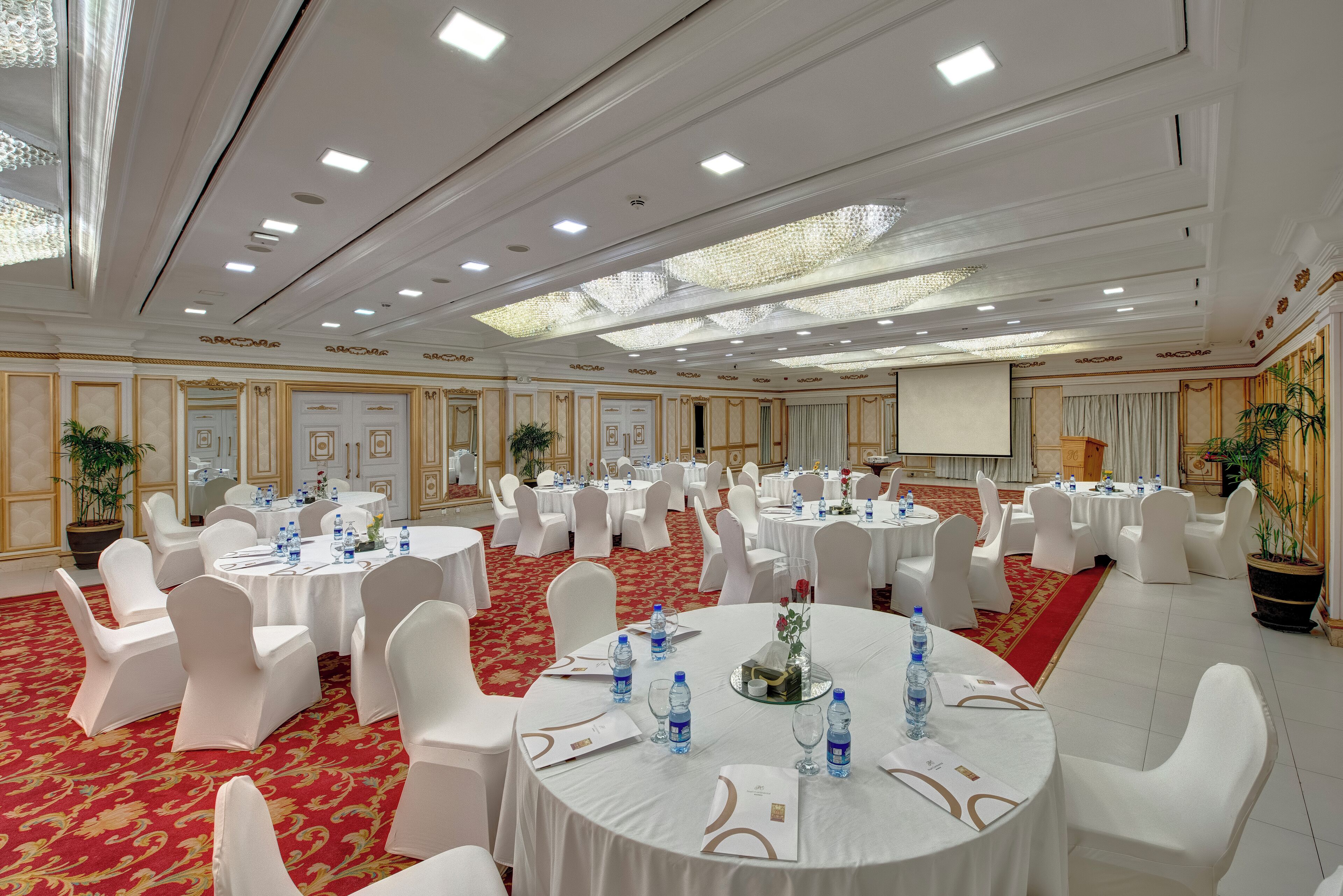 Une salle de banquet avec des tables rondes, des chaises blanches et des tapis rouges.