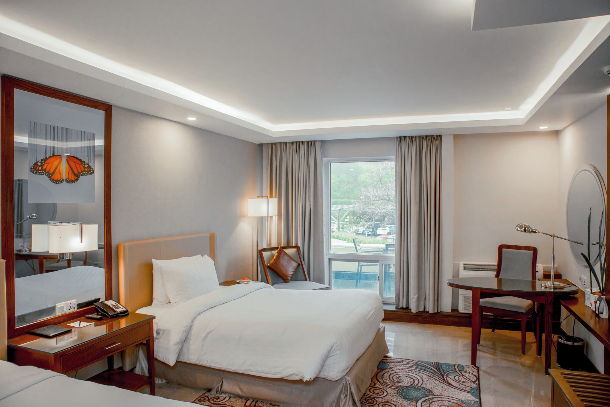 A hotel room with a large bed, a desk with a chair, a mirror, and a window with curtains.