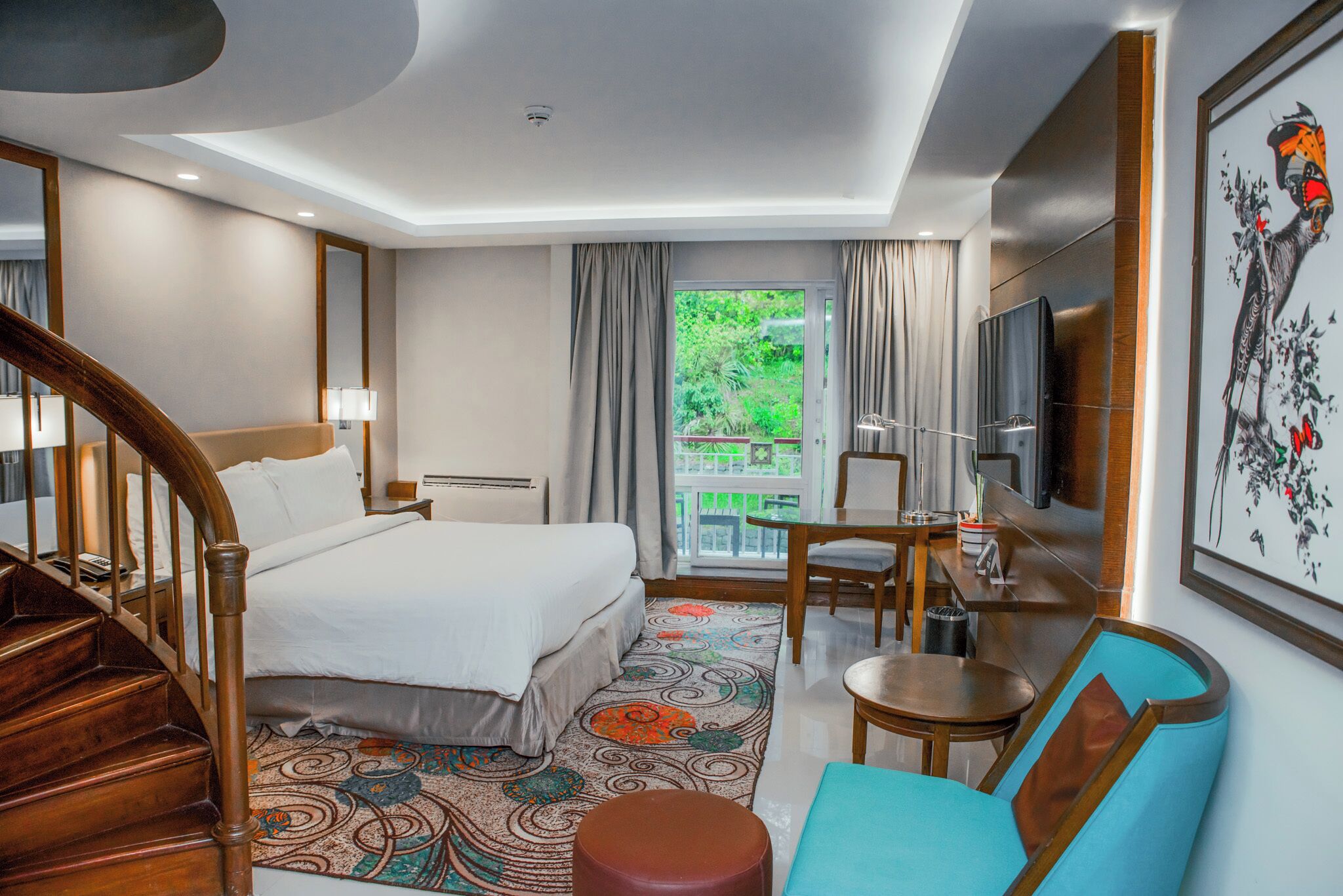 Habitación de hotel con cama, escritorio, silla, televisor, escalera y balcón con vista a zonas verdes.