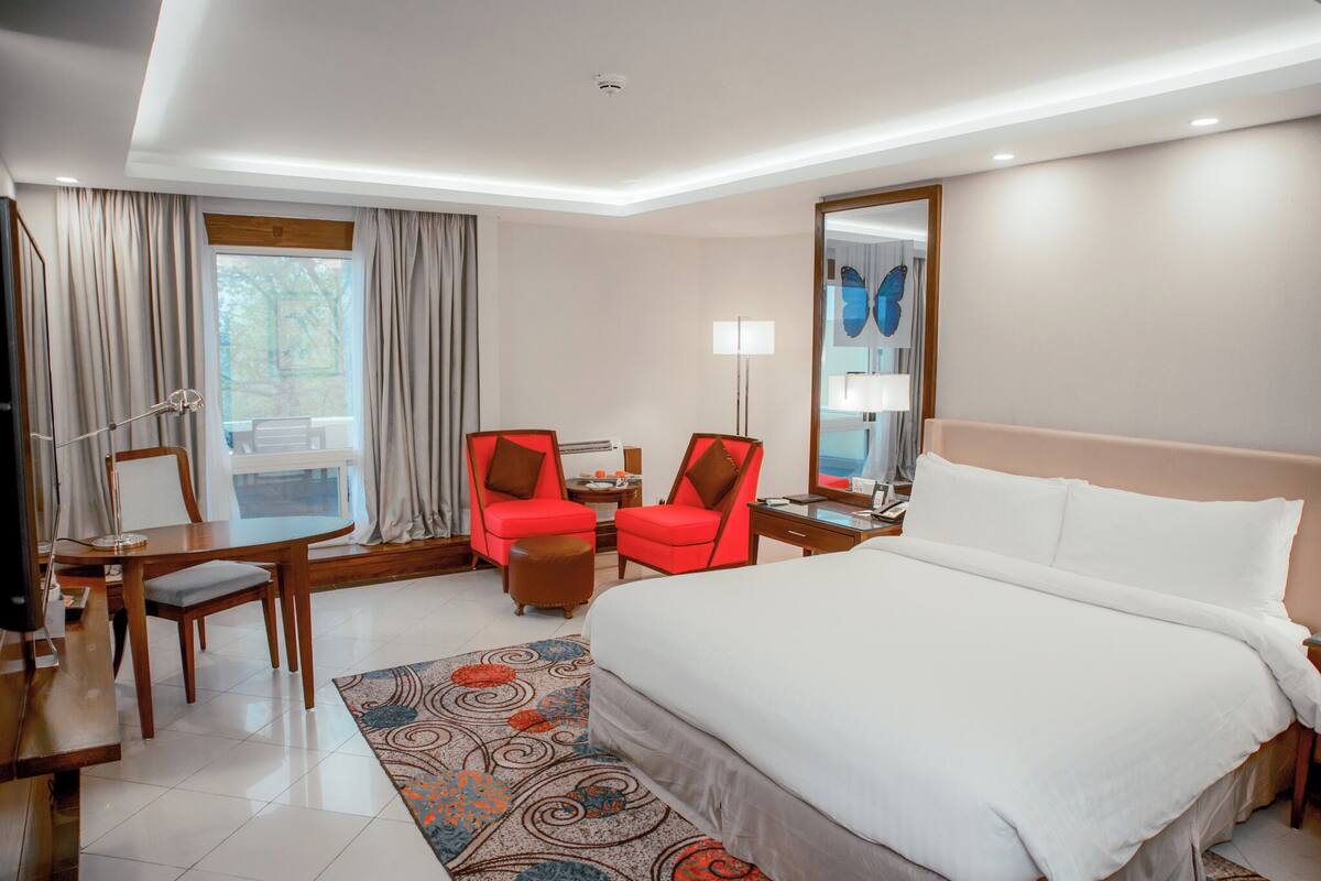 Une chambre d’hôtel avec un grand lit, deux fauteuils rouges, un bureau avec une lampe, un miroir et une vue sur l’extérieur.