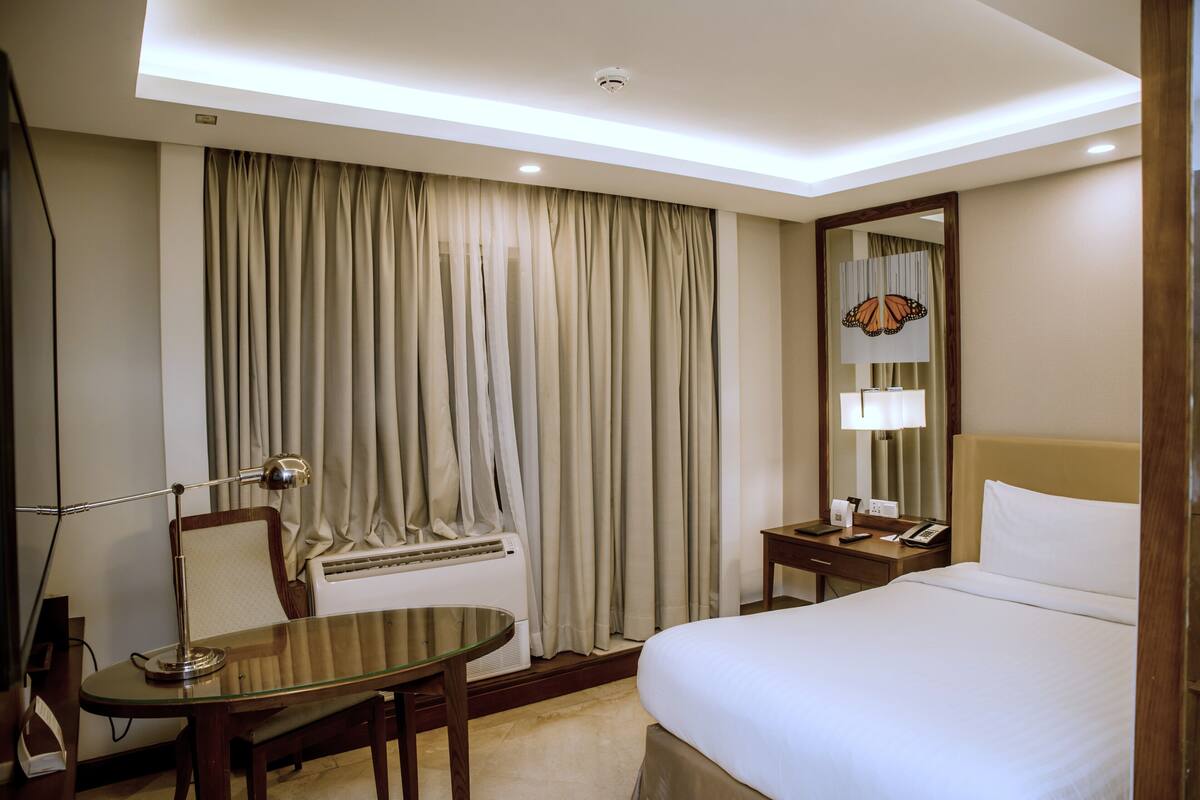 Une chambre d’hôtel équipée d’un lit, d’un bureau, d’une chaise, d’une lampe, d’un miroir et d’une fenêtre avec des rideaux.