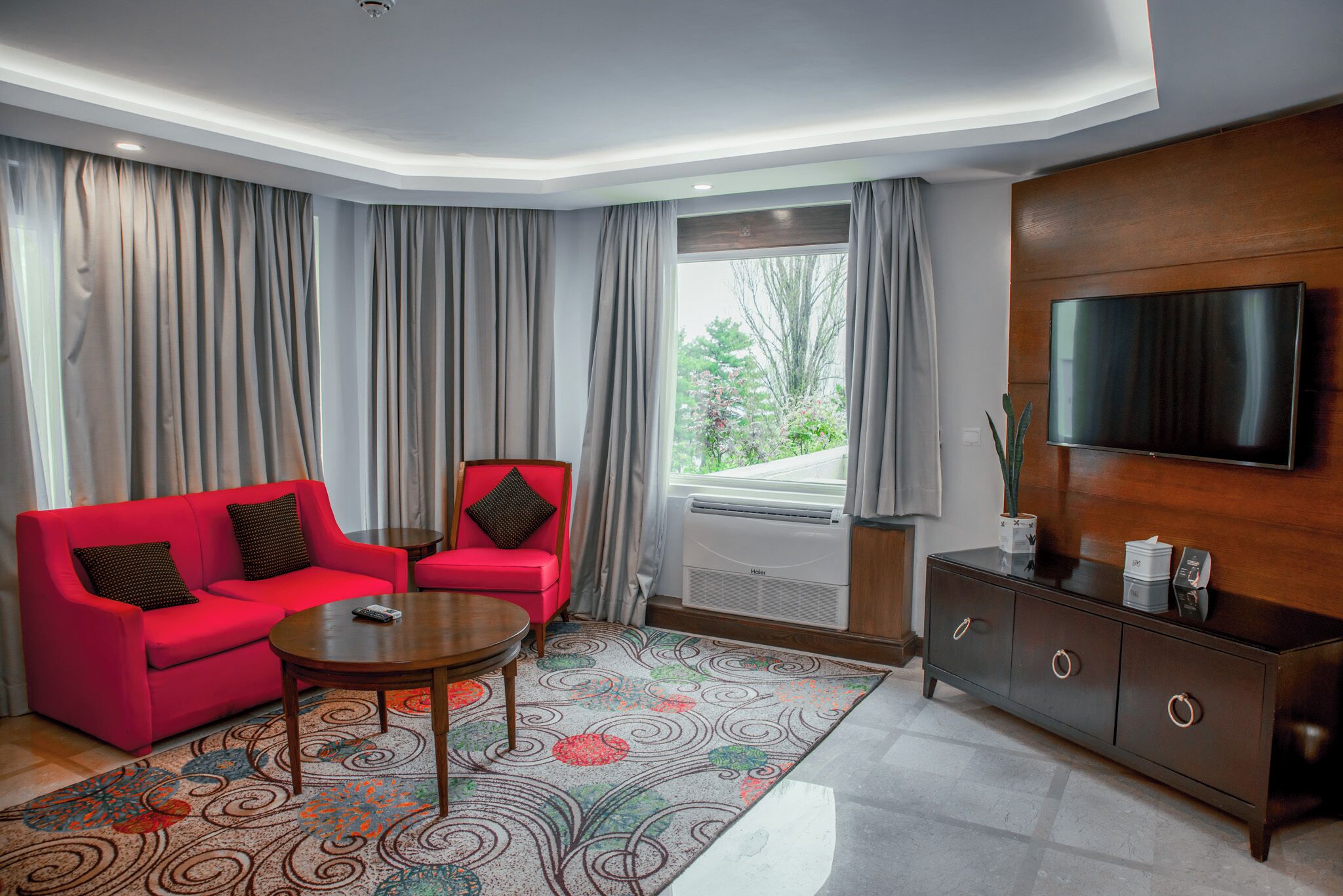 A modern hotel room with a red sofa, a small table, a TV, and a large window with curtains.