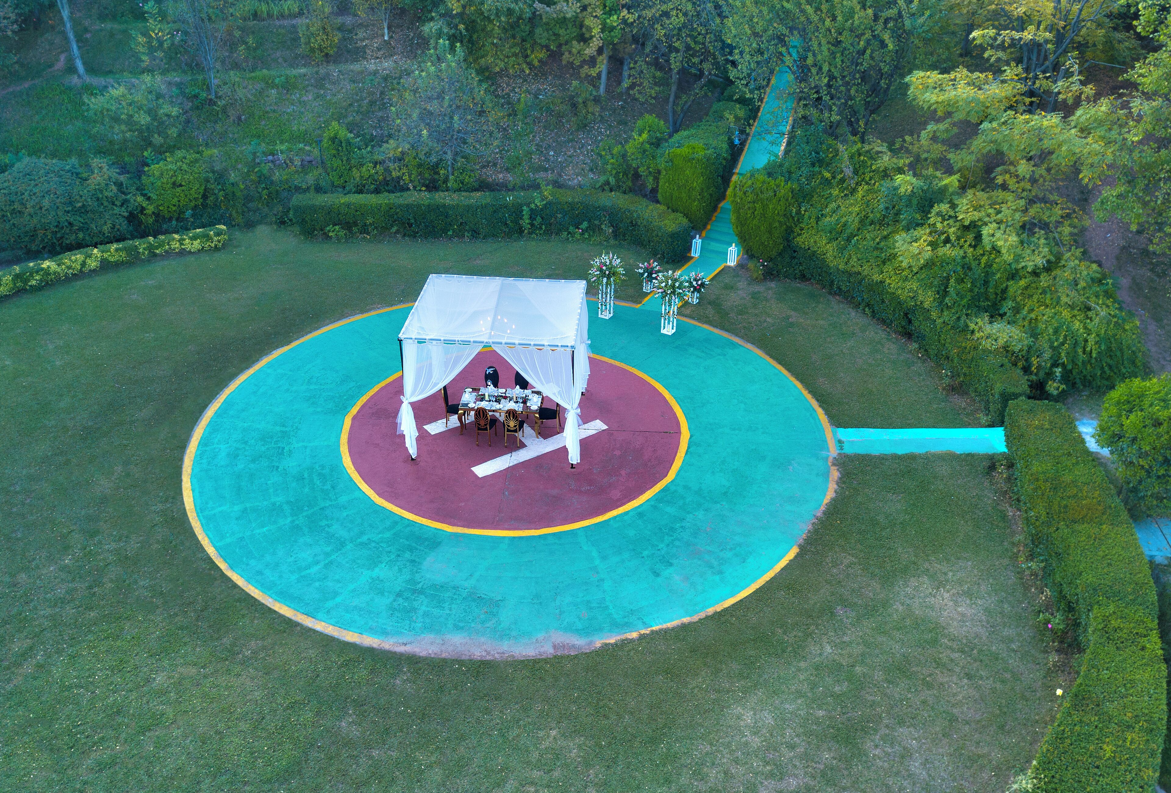 Vista aérea de uma área externa circular com uma tenda e mesas, cercada por um gramado verde e árvores.
