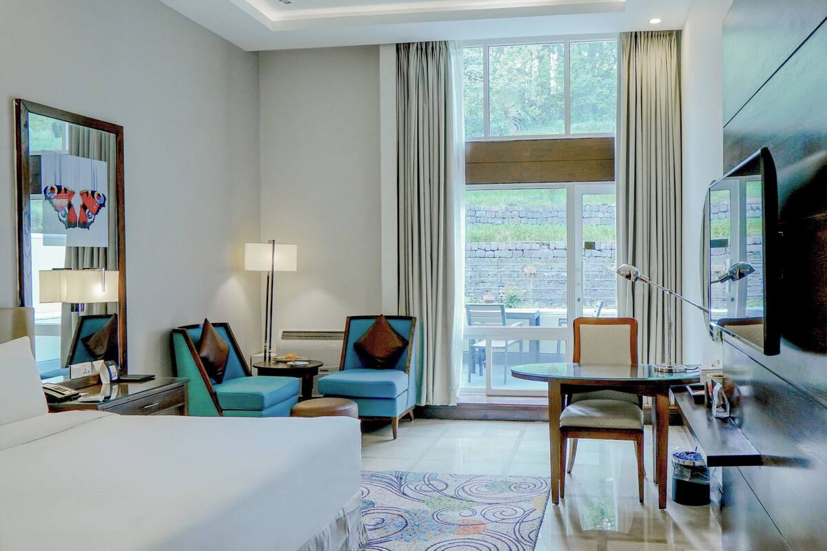 Une chambre d’hôtel avec un lit, deux fauteuils, une petite table, un téléviseur et une fenêtre avec des rideaux.
