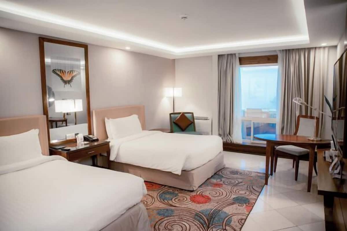 A hotel room with two beds, a desk, a chair, a lamp, a mirror, and a view of the outside.