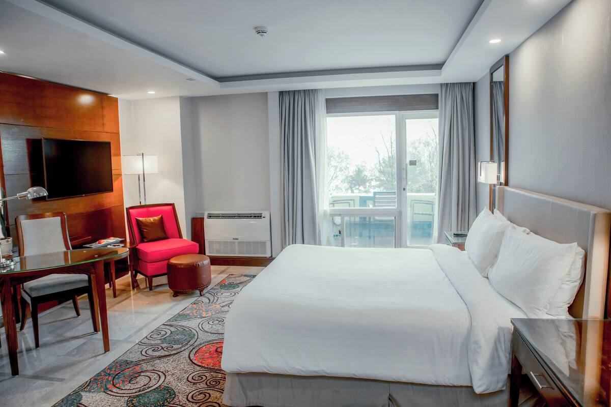 Une chambre d’hôtel avec un grand lit, un bureau avec une chaise, un téléviseur fixé au mur et un balcon avec des rideaux.