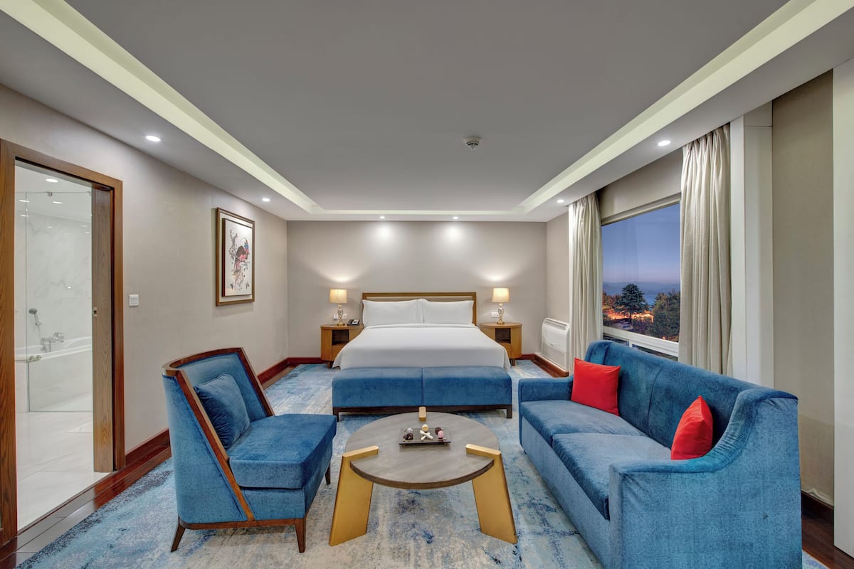 Une chambre d’hôtel avec un grand lit, deux canapés bleus, une table basse ronde et une vue sur le paysage urbain par la fenêtre.