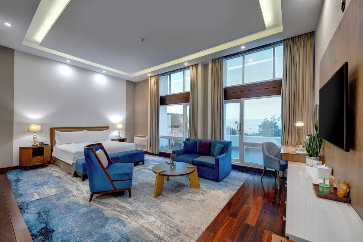 Une chambre d’hôtel avec un lit, des fauteuils bleus, un canapé, une table basse, une télévision et un balcon avec vue.