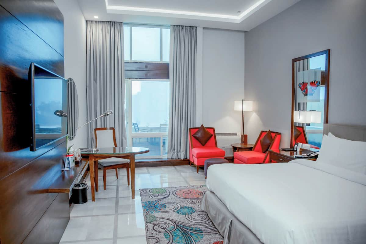 Une chambre d’hôtel équipée d’un lit, d’une télévision, d’un bureau, de deux chaises, d’une lampe et d’une fenêtre avec des rideaux.