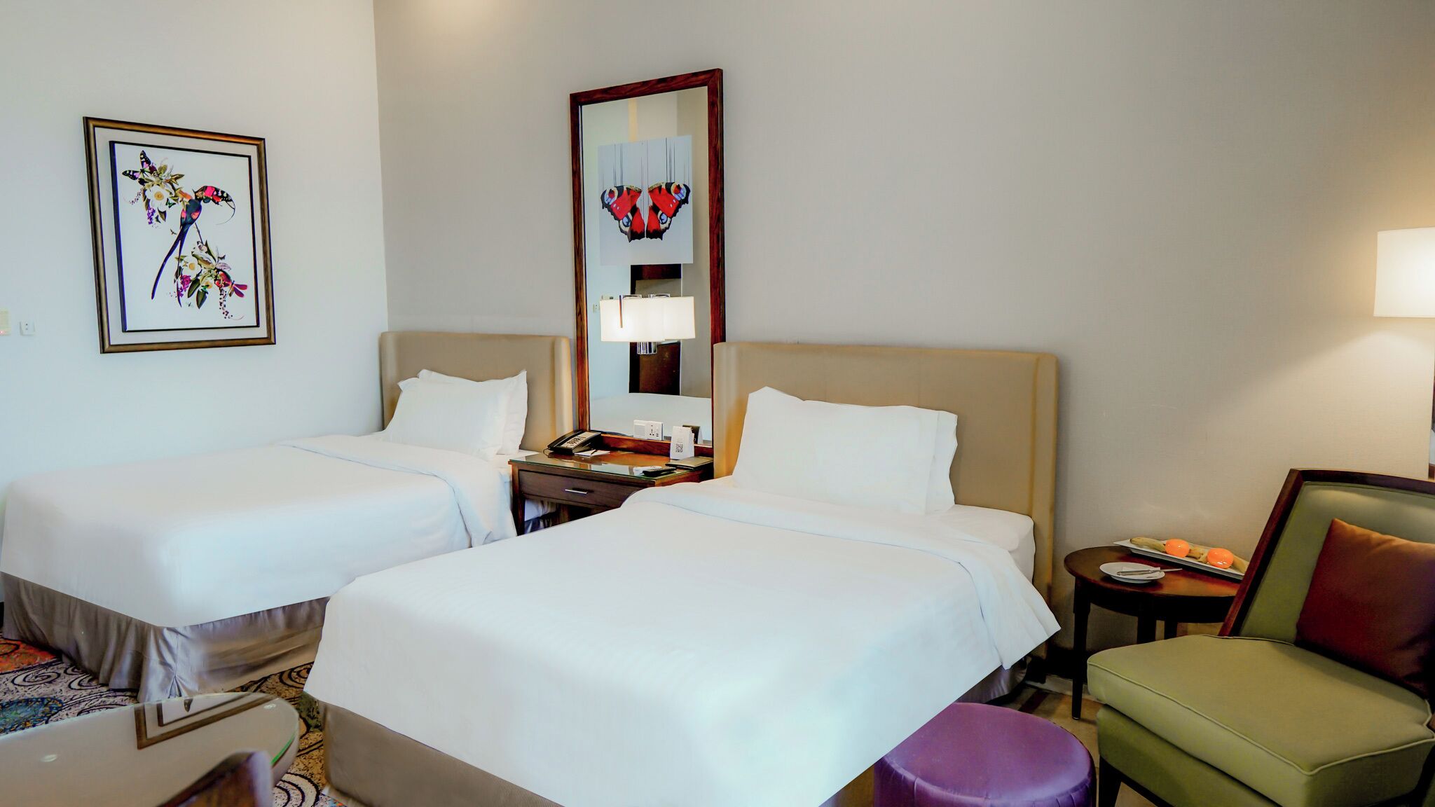 A hotel room with two beds, a bedside table with a lamp, a chair, and artwork on the walls.