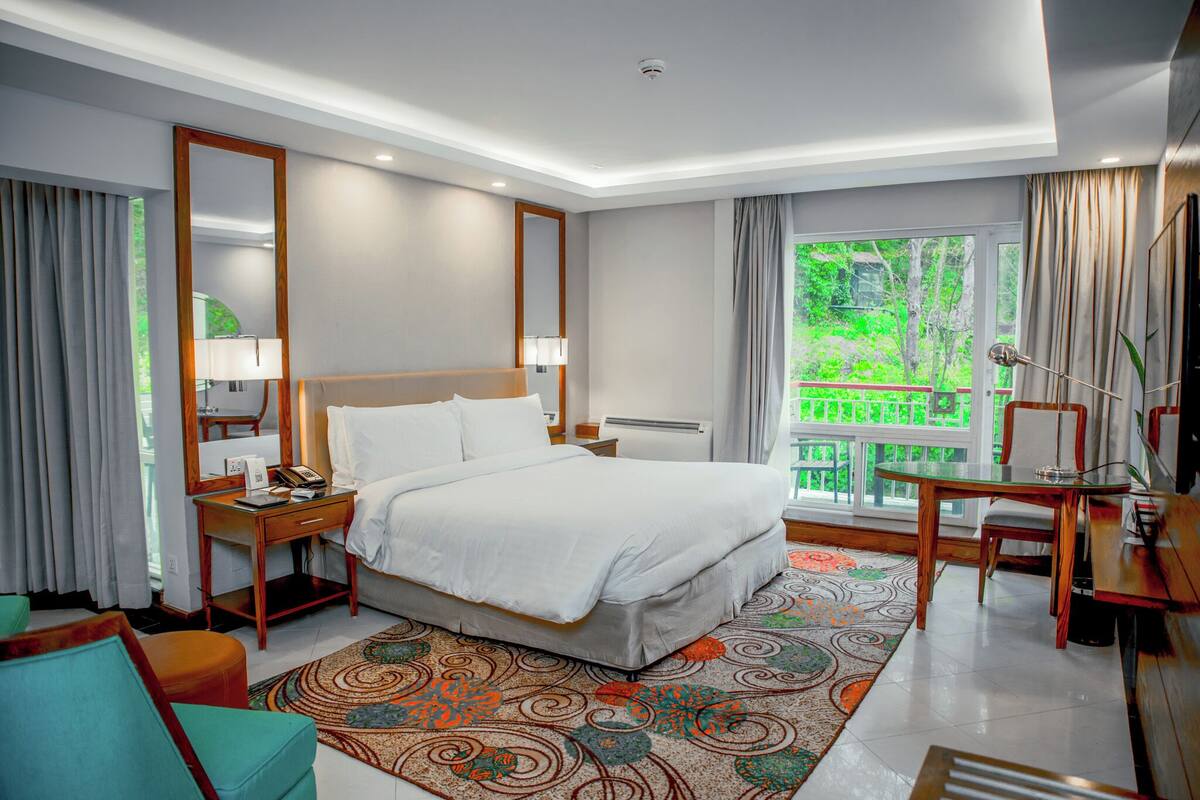Une chambre d’hôtel équipée d’un lit, d’un bureau, d’une chaise, d’une table de chevet, d’une lampe, d’un miroir et d’une fenêtre donnant sur un paysage verdoyant.
