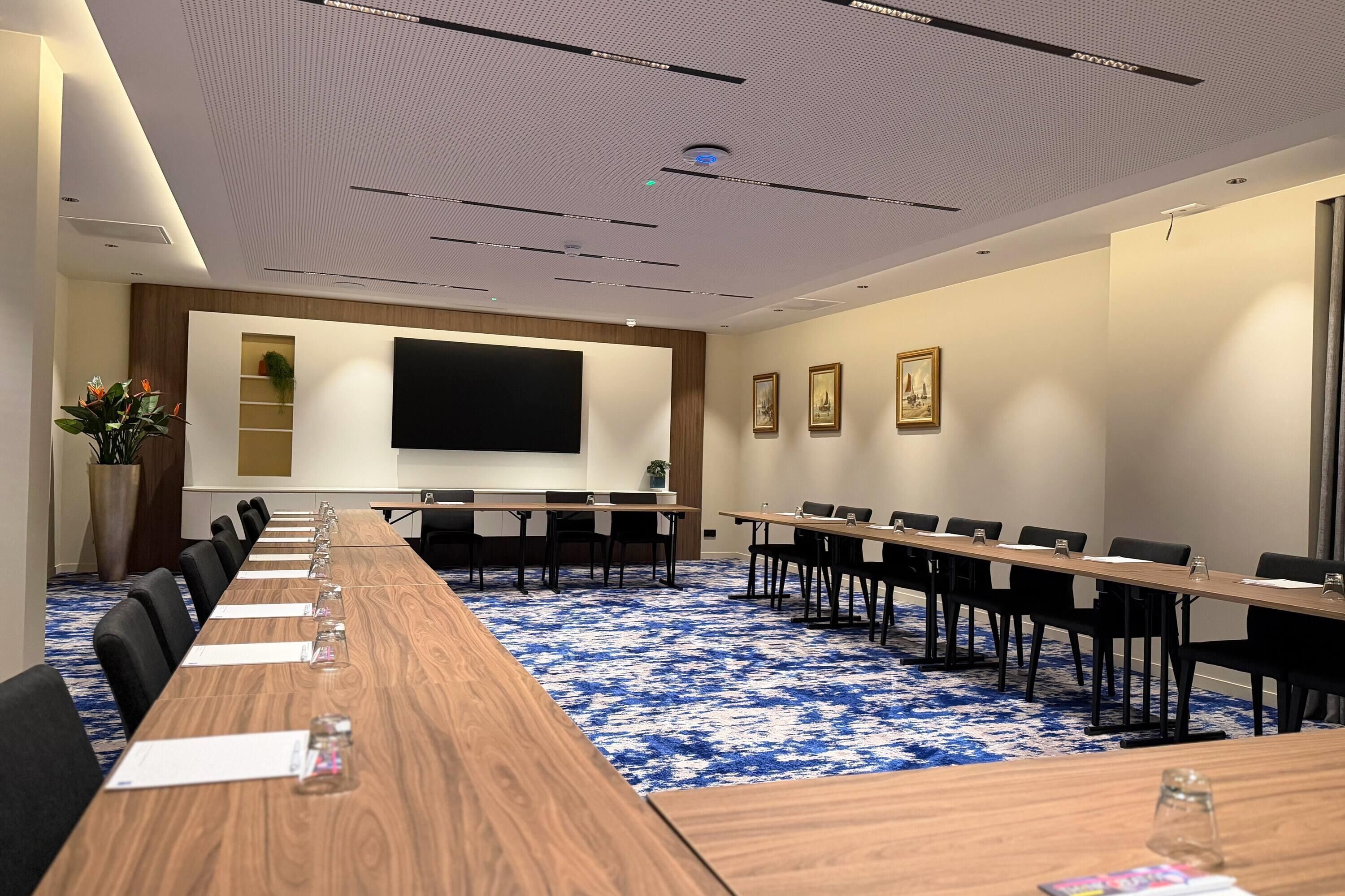 Un salón de conferencias con una larga mesa, sillas, una pantalla de proyector y una alfombra azul estampada.