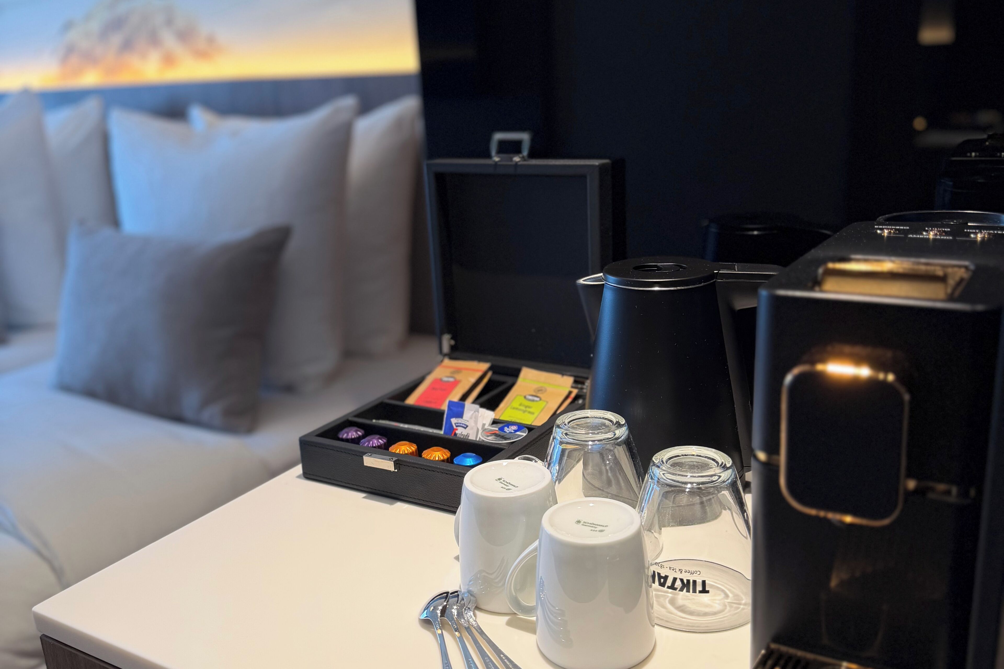 A hotel room with a coffee machine, a tray of coffee pods, a glass jar, and a white mug on a table.
