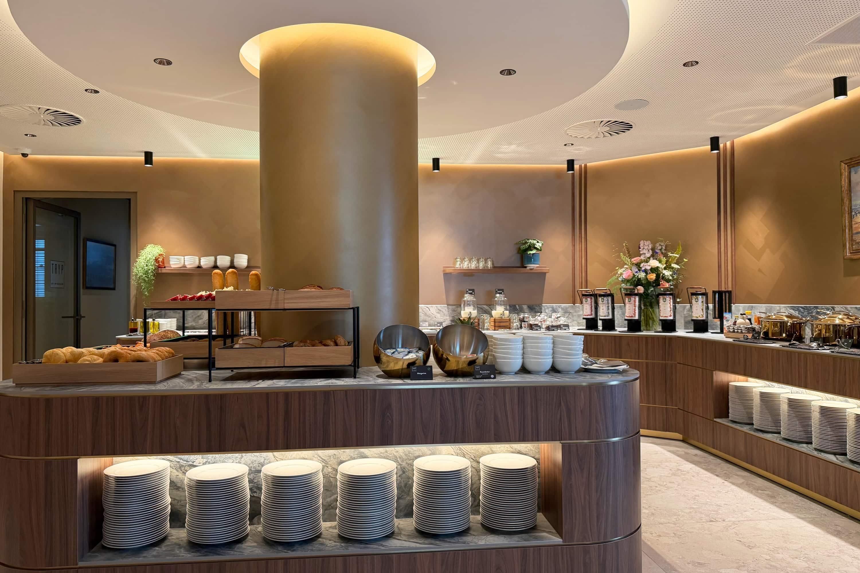 A modern hotel lobby with a central reception desk, a buffet area with various food items, and a display of stacked plates.