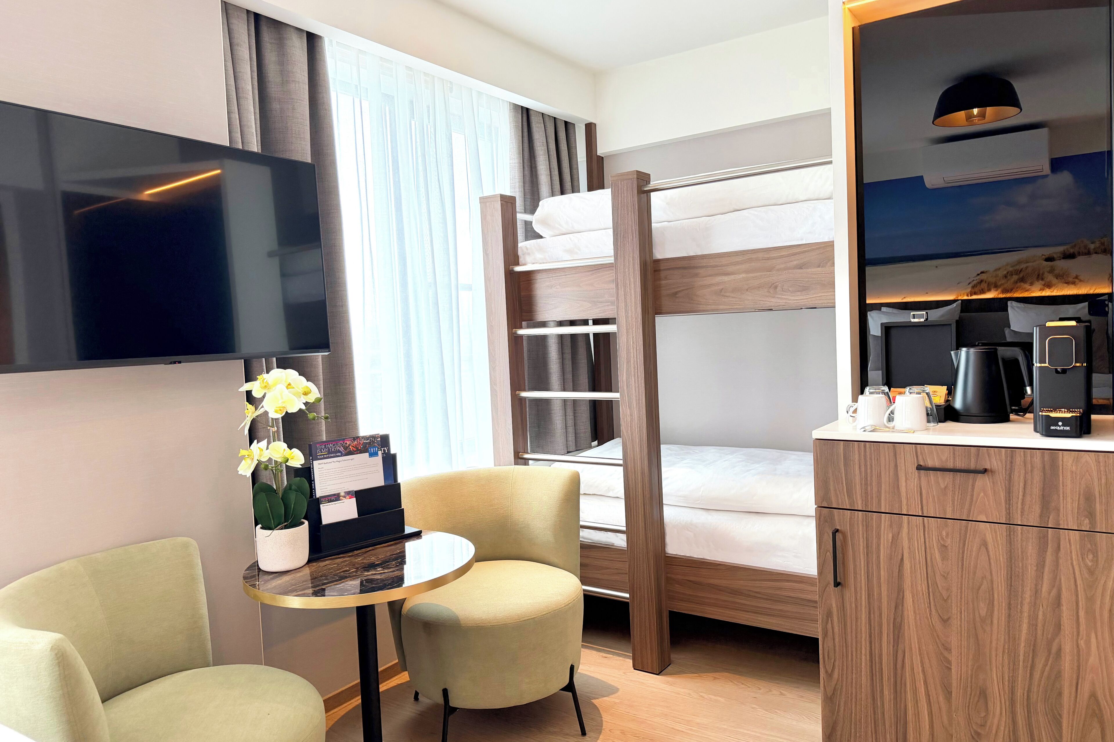 A hotel room with a bunk bed, a flat-screen TV, a small round table with a chair, a potted plant, and a coffee maker.