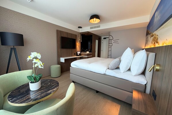 Una habitación de hotel moderna con una cama, un escritorio, una silla, una mesa redonda con una planta y un cuadro en la pared.