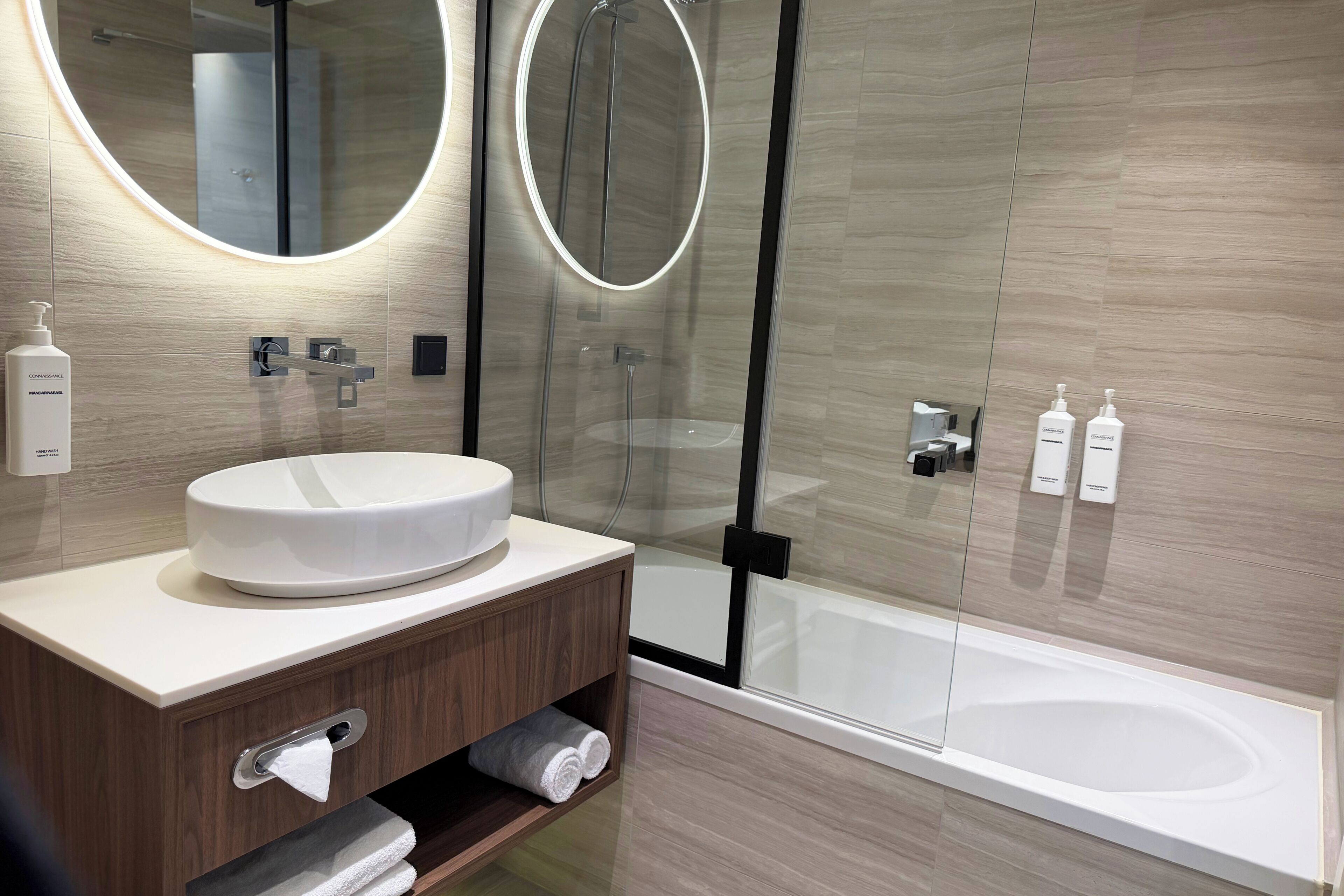 Un baño moderno con un espejo redondo, un lavabo, ducha con mampara de cristal y bañera.