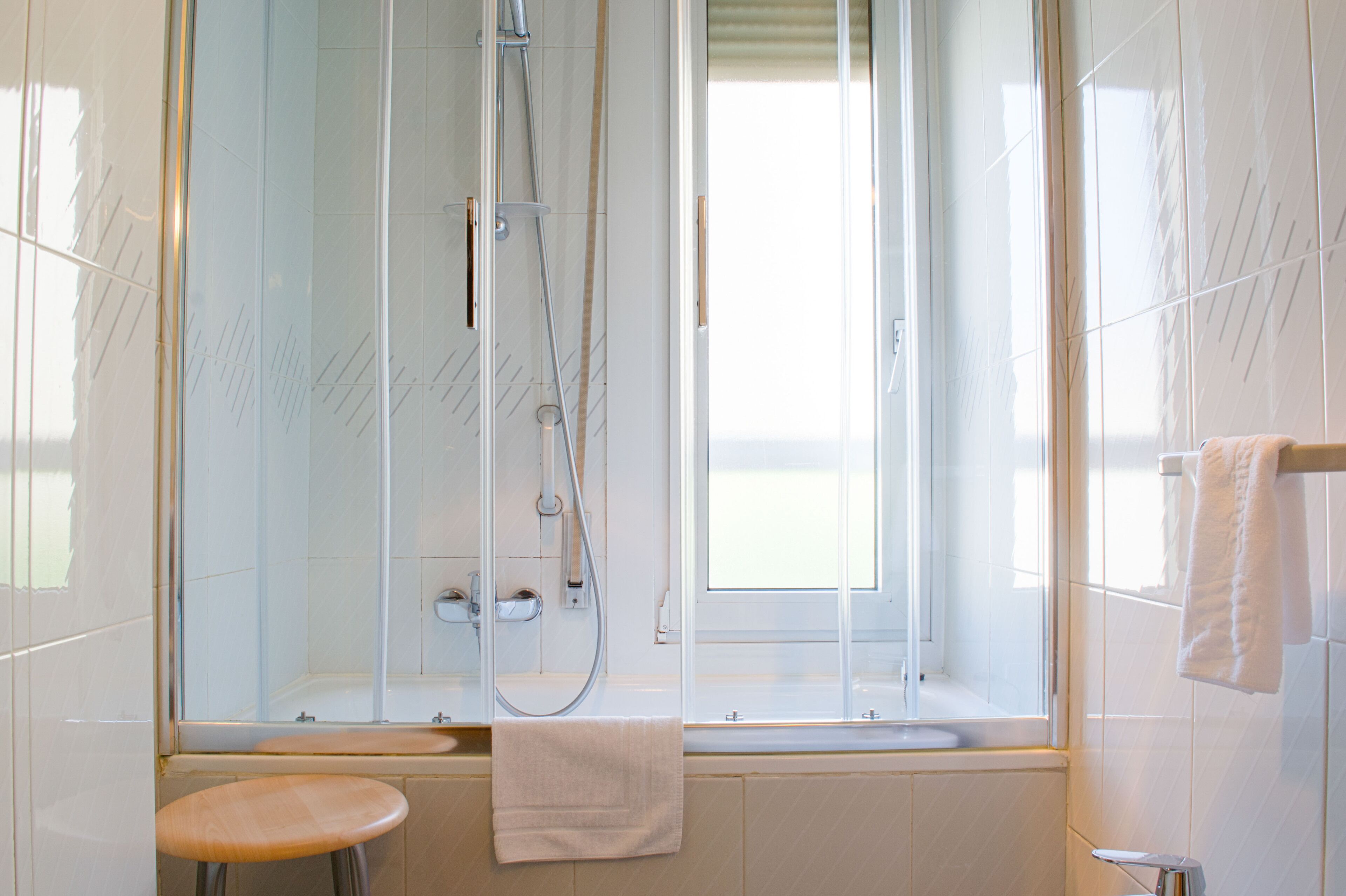 A modern bathroom with a glass-enclosed shower, a window, and a wooden stool.