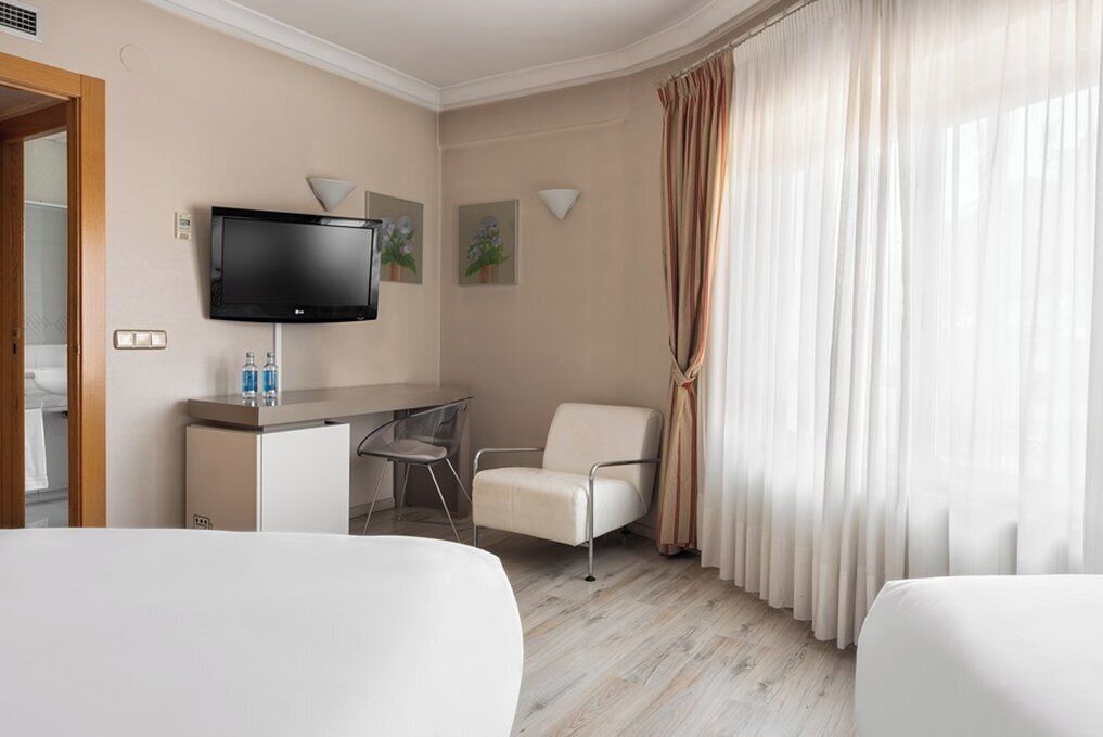Una habitación de hotel con cama, escritorio, silla, televisor y ventana con cortinas.