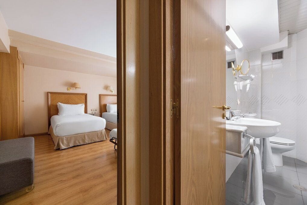 Una habitación de hotel con piso de madera, cama con ropa de cama blanca, mesita de noche con lámpara y baño con lavamanos e inodoro.