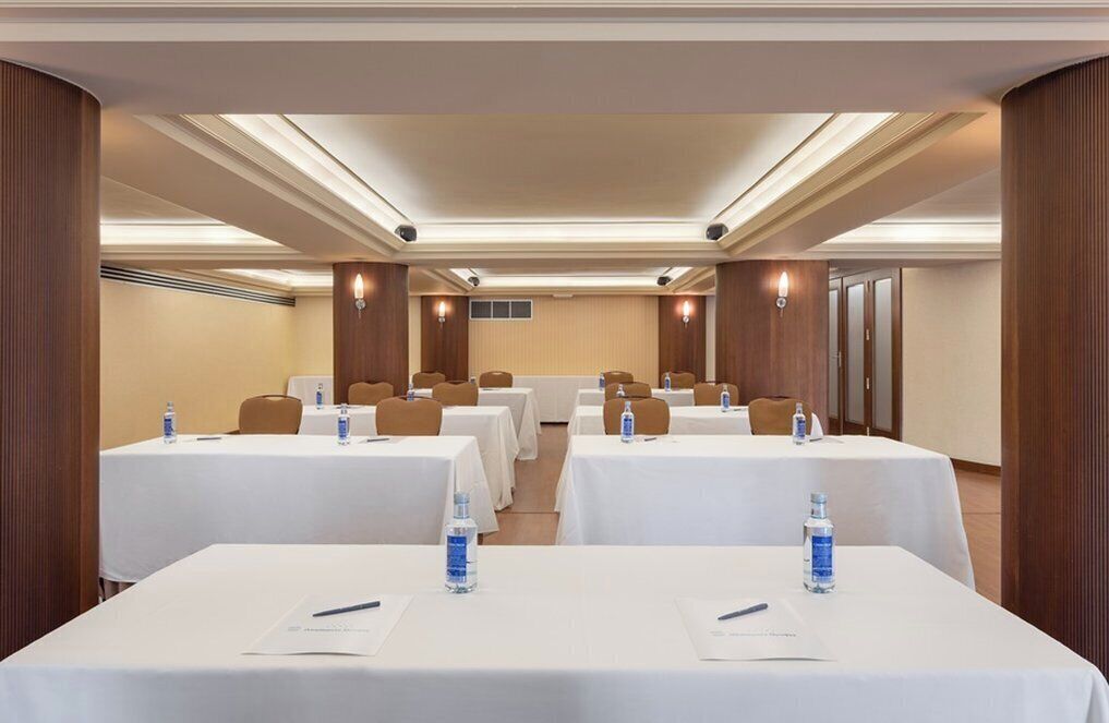 Une salle de conférence dotée de plusieurs tables, chacune recouverte d’une nappe blanche, avec des bouteilles d’eau et des stylos.