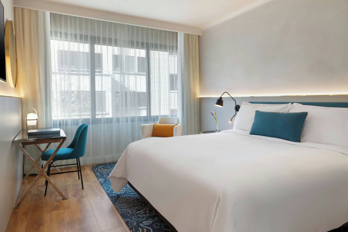 A hotel room with a large bed, a desk with a chair, a window with sheer curtains, and a bedside table with a lamp.