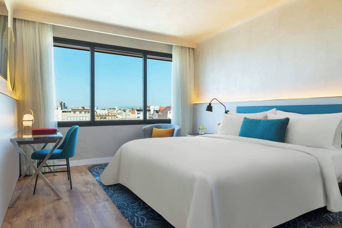 A hotel room with a large bed, a desk with a chair, a view of the city through the window, and a nightstand with a lamp.