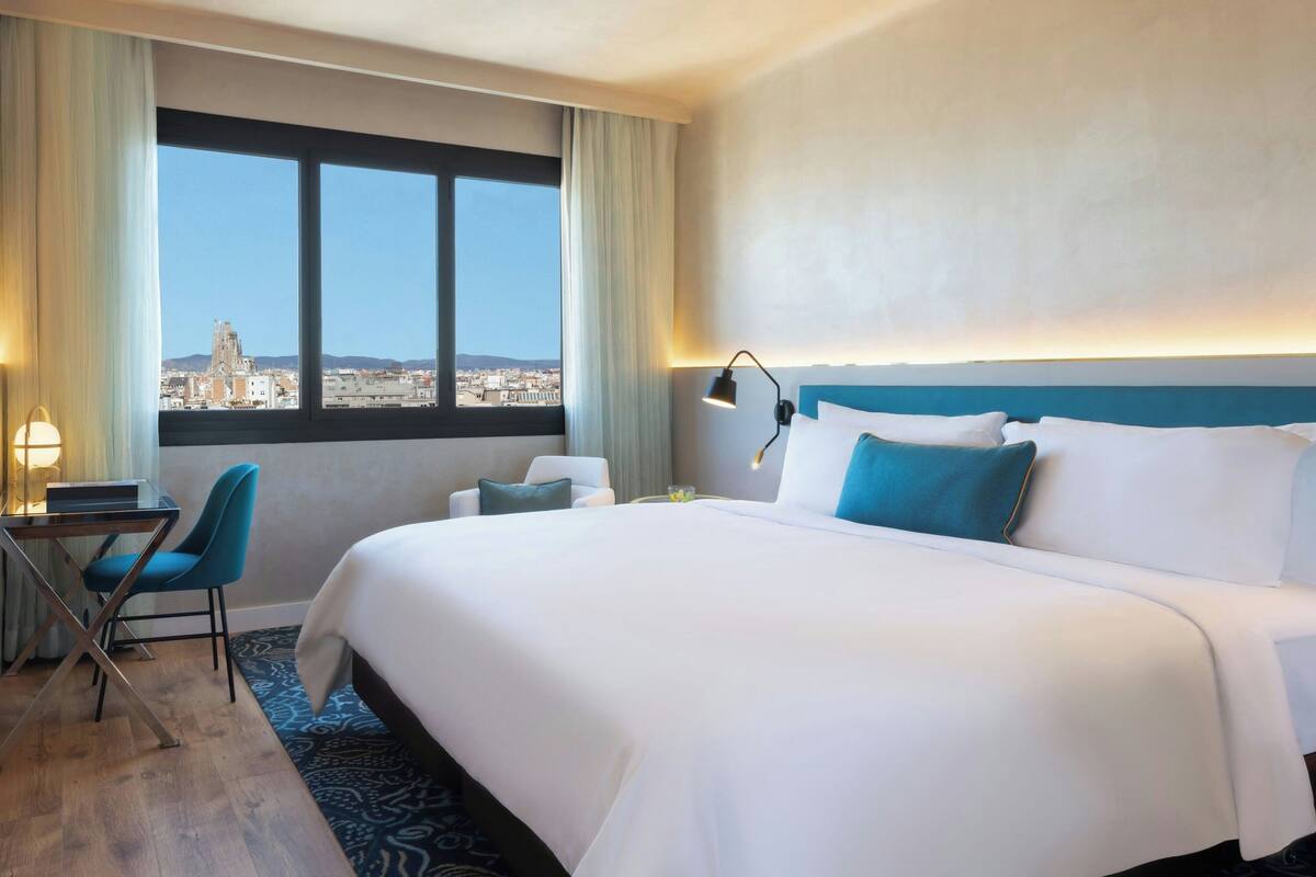 A hotel room with a large bed, a desk, a chair, and a view of a cityscape through a window.