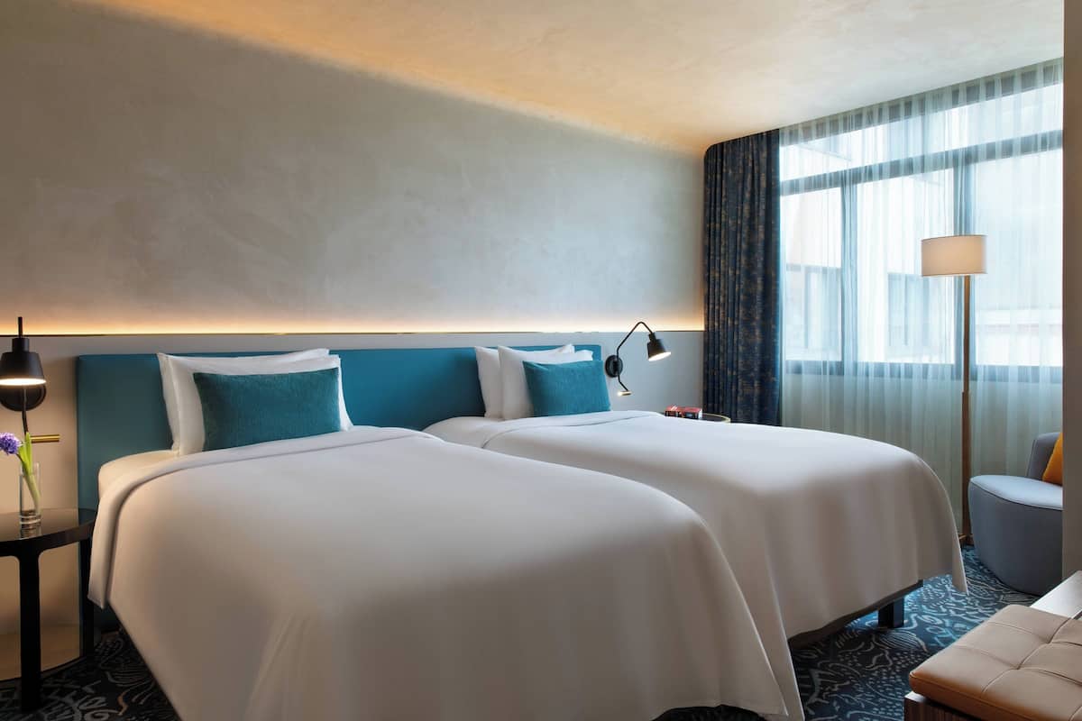 A hotel room with two beds, a nightstand, a lamp, and a view of the outdoors.