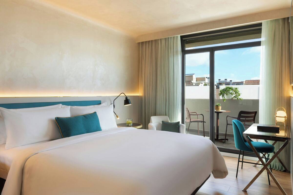A hotel room with a large bed, a desk, and a chair. There is a window with curtains and a view of buildings outside.