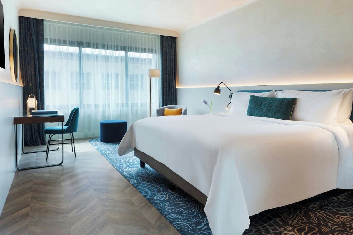 A hotel room with a large bed, a desk with a chair, a lamp, and a view of the city through large windows.