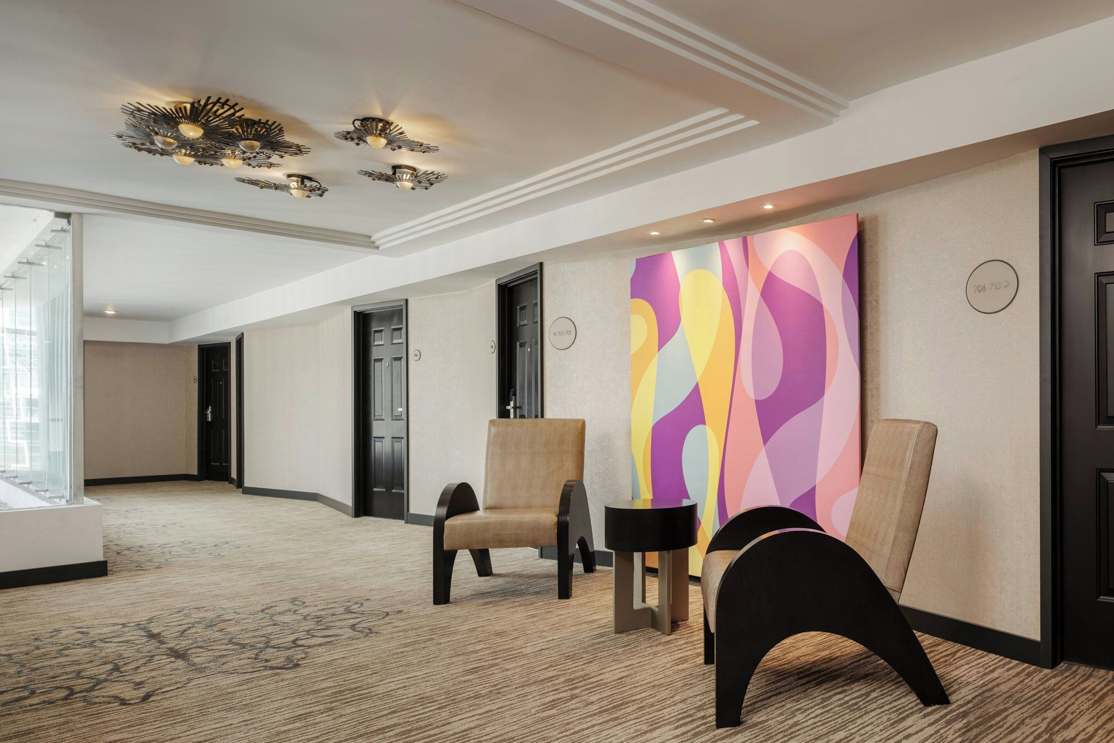 Een hotellobby met vloerbedekking, twee stoelen, een kleurrijk abstract schilderij en een plafondlamp.