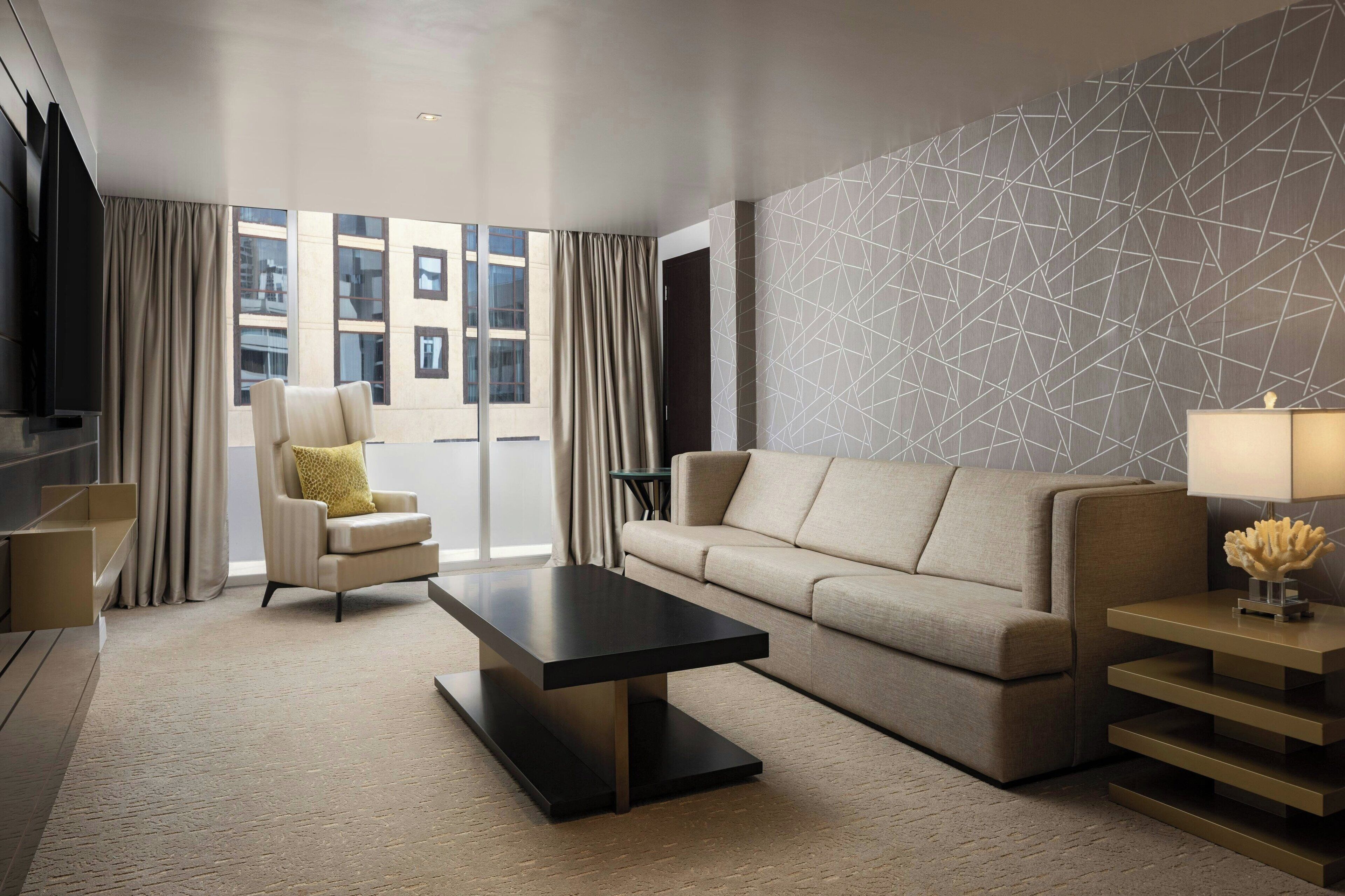 A modern living room with a sofa, armchair, and coffee table, large windows with curtains, and a geometric-patterned wall.