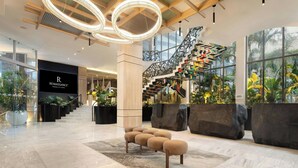 Un vestíbulo moderno con una escalera, suelo de mármol y grandes ventanales con plantas tropicales.