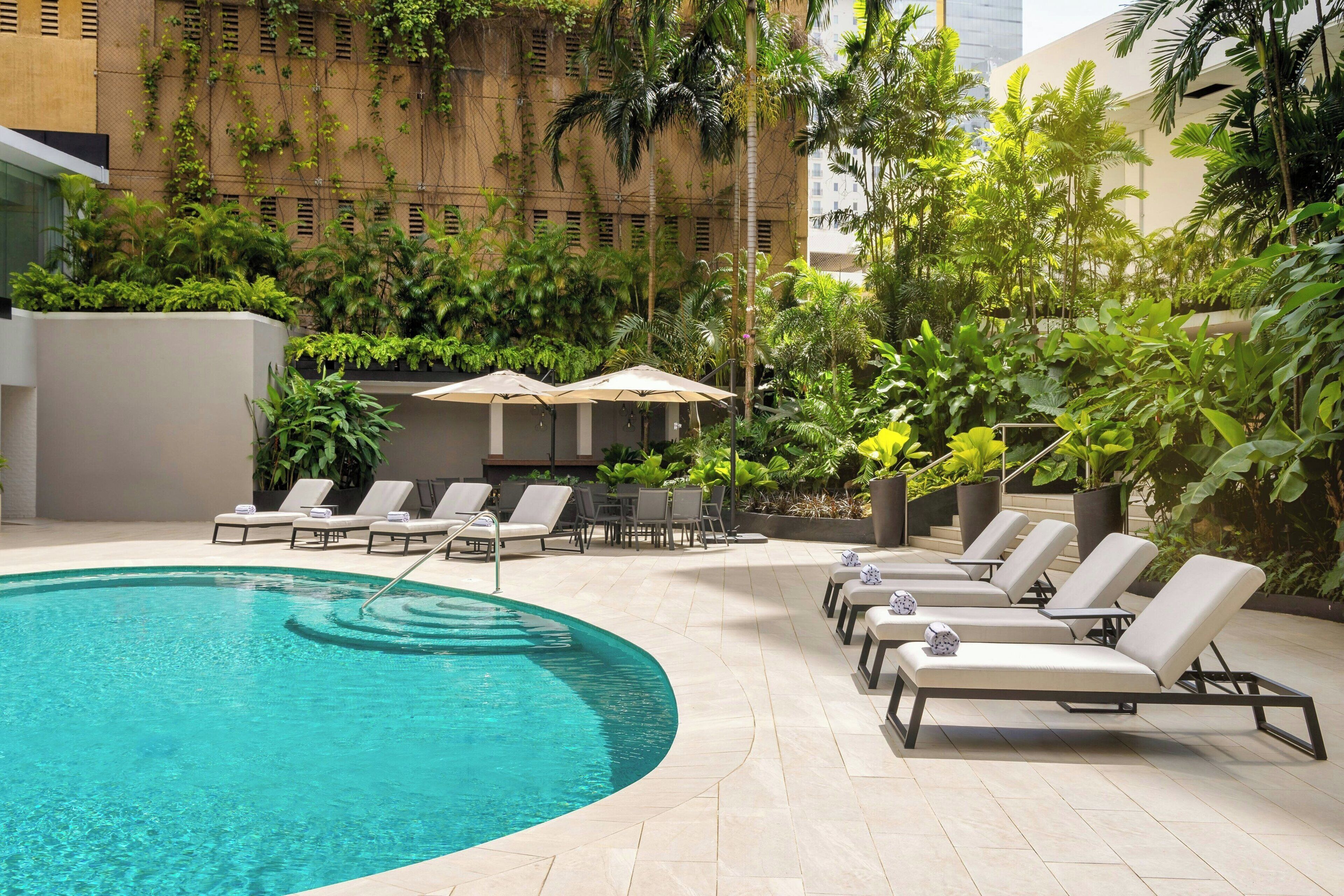 Un área junto a la piscina con sillones de descanso y sombrillas, rodeada de exuberante vegetación y una fachada de edificio moderna.