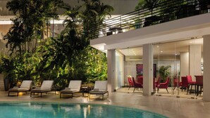 Una piscina interior con sillones de descanso y sillas rojas, rodeada de exuberante vegetación y un diseño interior moderno.
