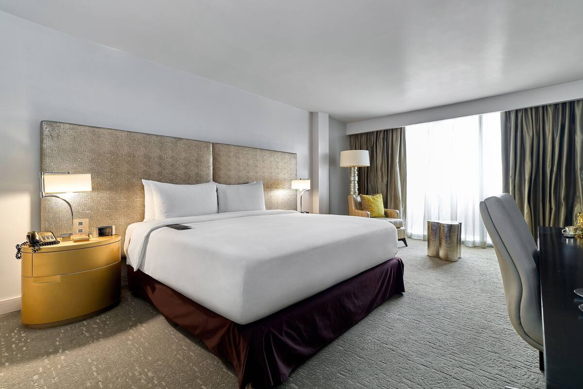 Habitación de hotel con una cama grande, una mesita de noche con lámpara, una silla y vistas al exterior.