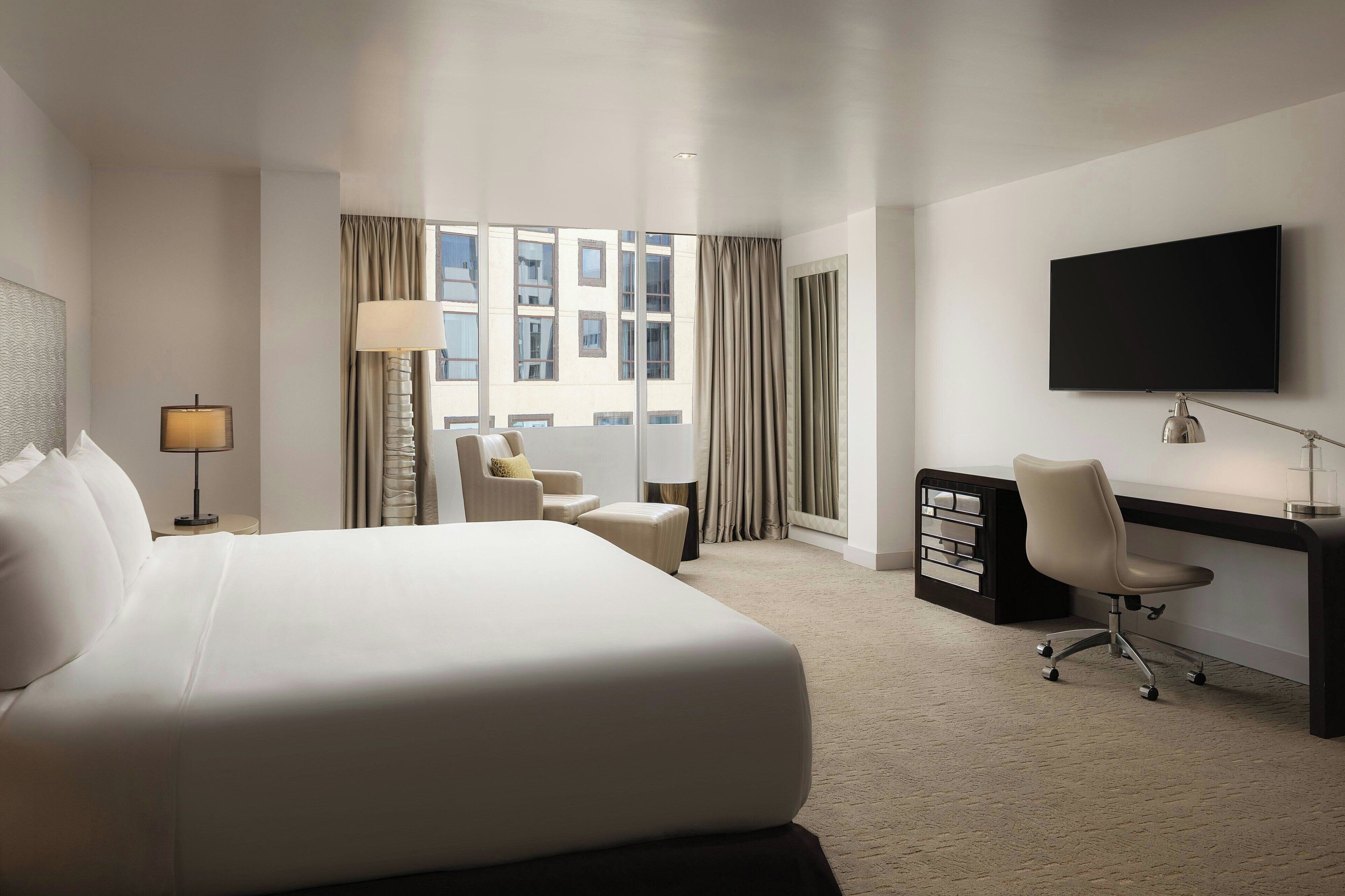 A hotel room with a large bed, a desk, a chair, a TV, and a window with curtains.
