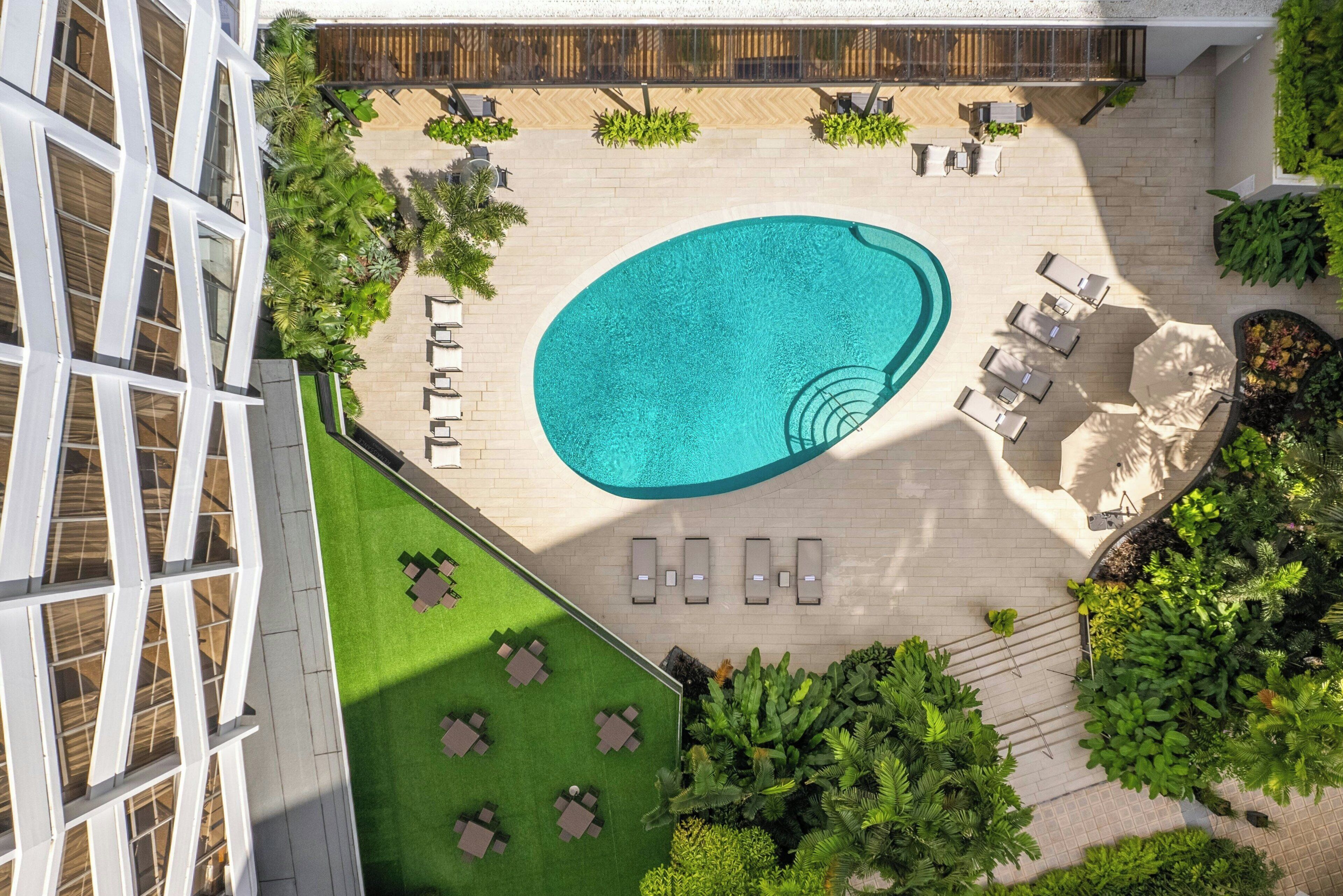 An aerial view of a rooftop pool area with a swimming pool, sun loungers, and surrounding greenery.