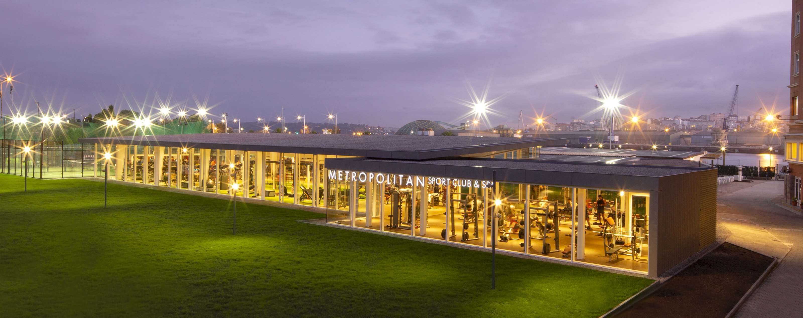 A modern sports club with a glass facade, outdoor lighting, and a view of the cityscape in the background.