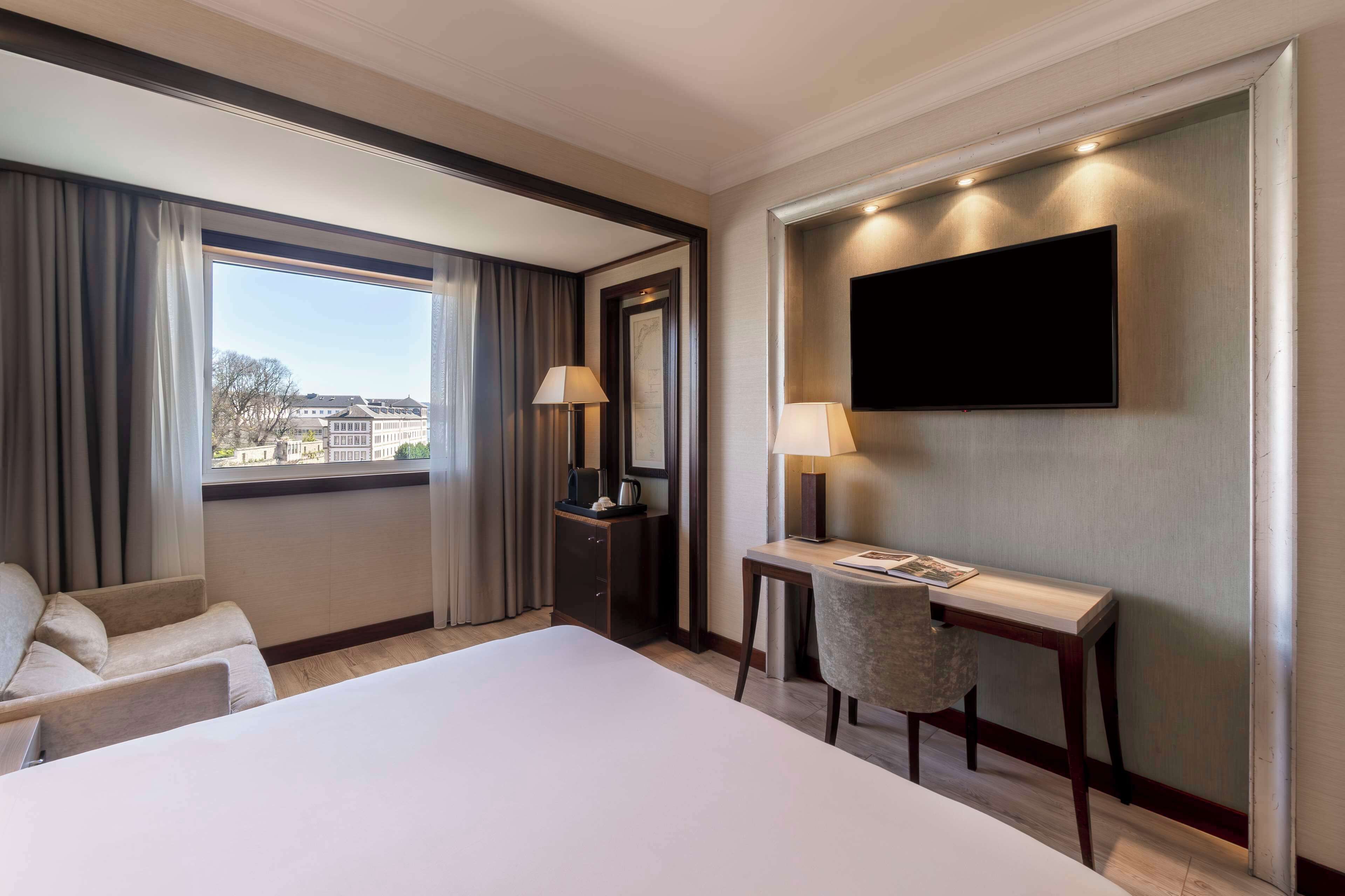 Une chambre d’hôtel avec un grand lit, un bureau avec une chaise, un téléviseur à écran plat fixé au mur et une fenêtre avec des rideaux.