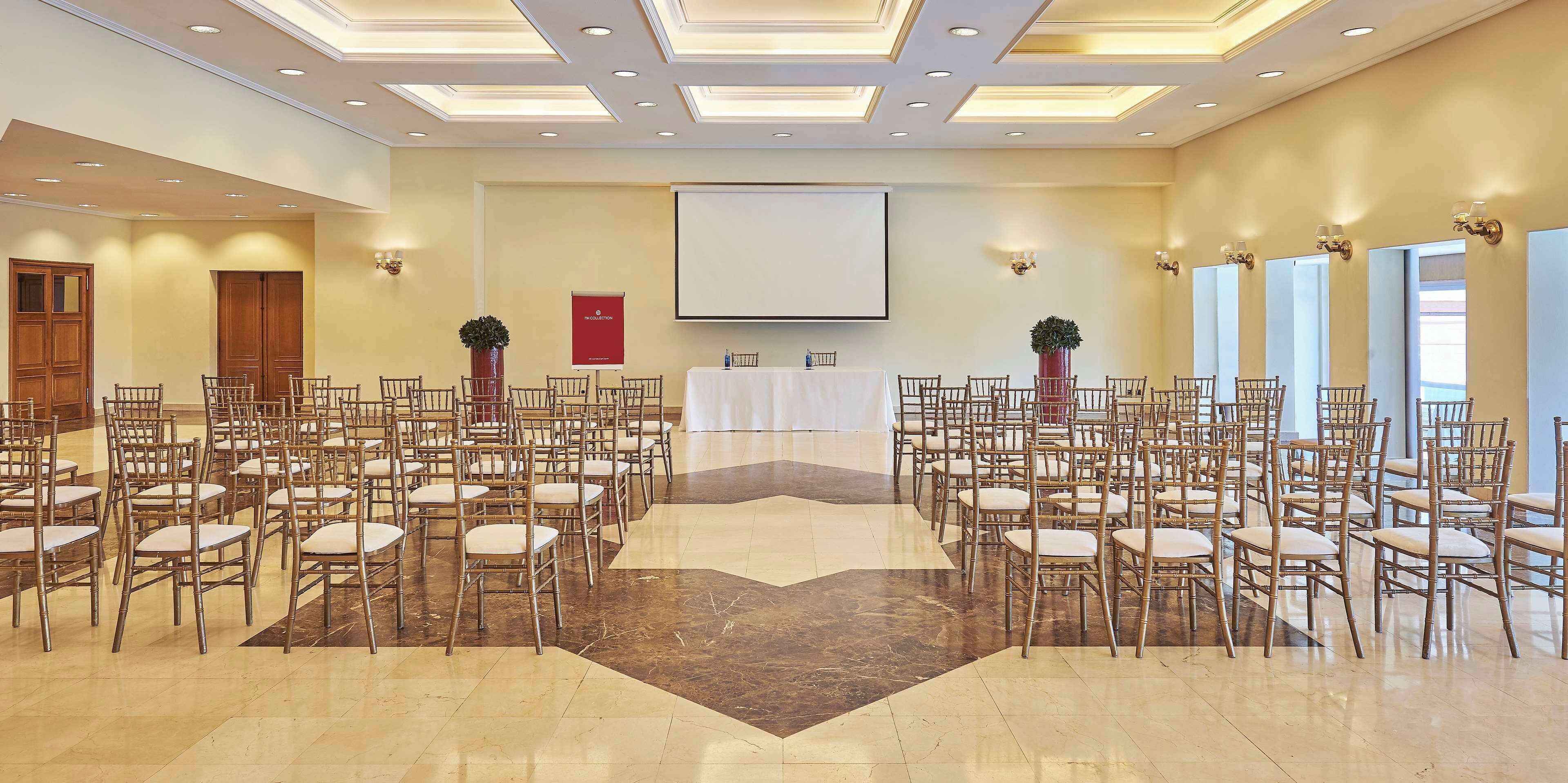 Un salón de conferencias con filas de sillas, un atril, una pantalla de proyector y un escenario.
